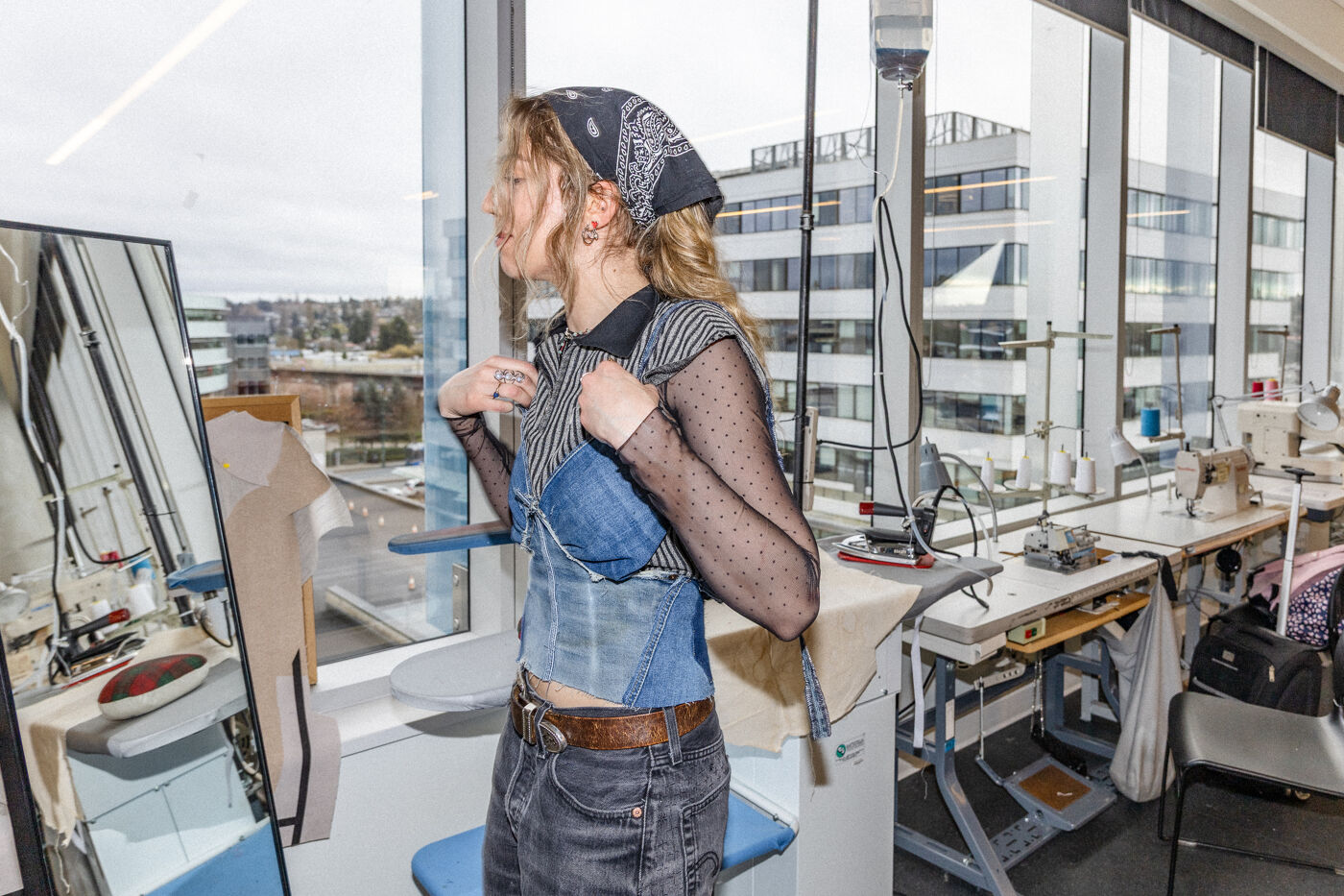 A young woman wearing a bandana tries on a denim corset in front of a mirror in a sewing workshop with sewing machines and irons in the background.