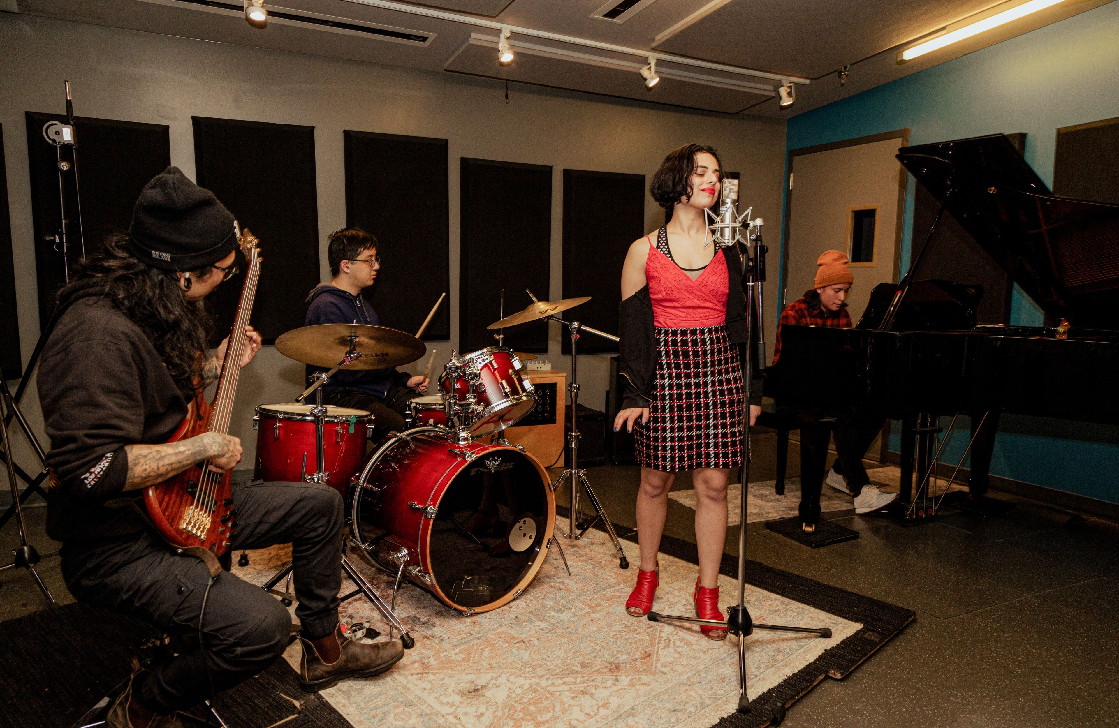 A band with vocalist, drummer, bassist, and pianist performing together in a studio.