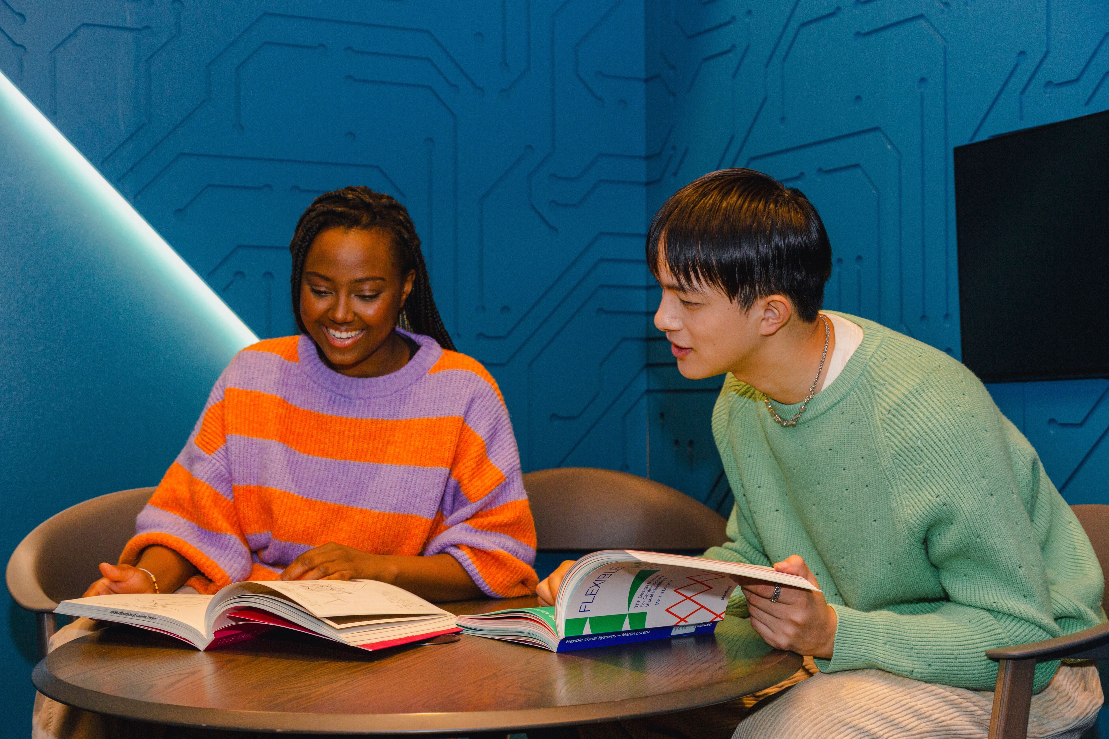 Two students, one in a striped sweater, review a book together in a vibrant study space.