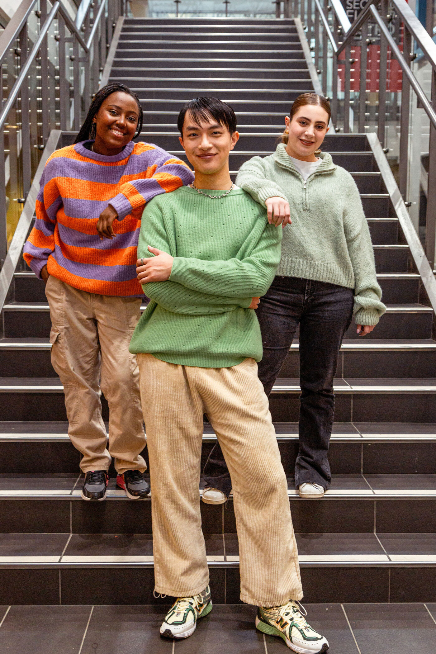Three students of diverse backgrounds posing on staircase, sporting casual sweaters and smiles, embodying campus life diversity.