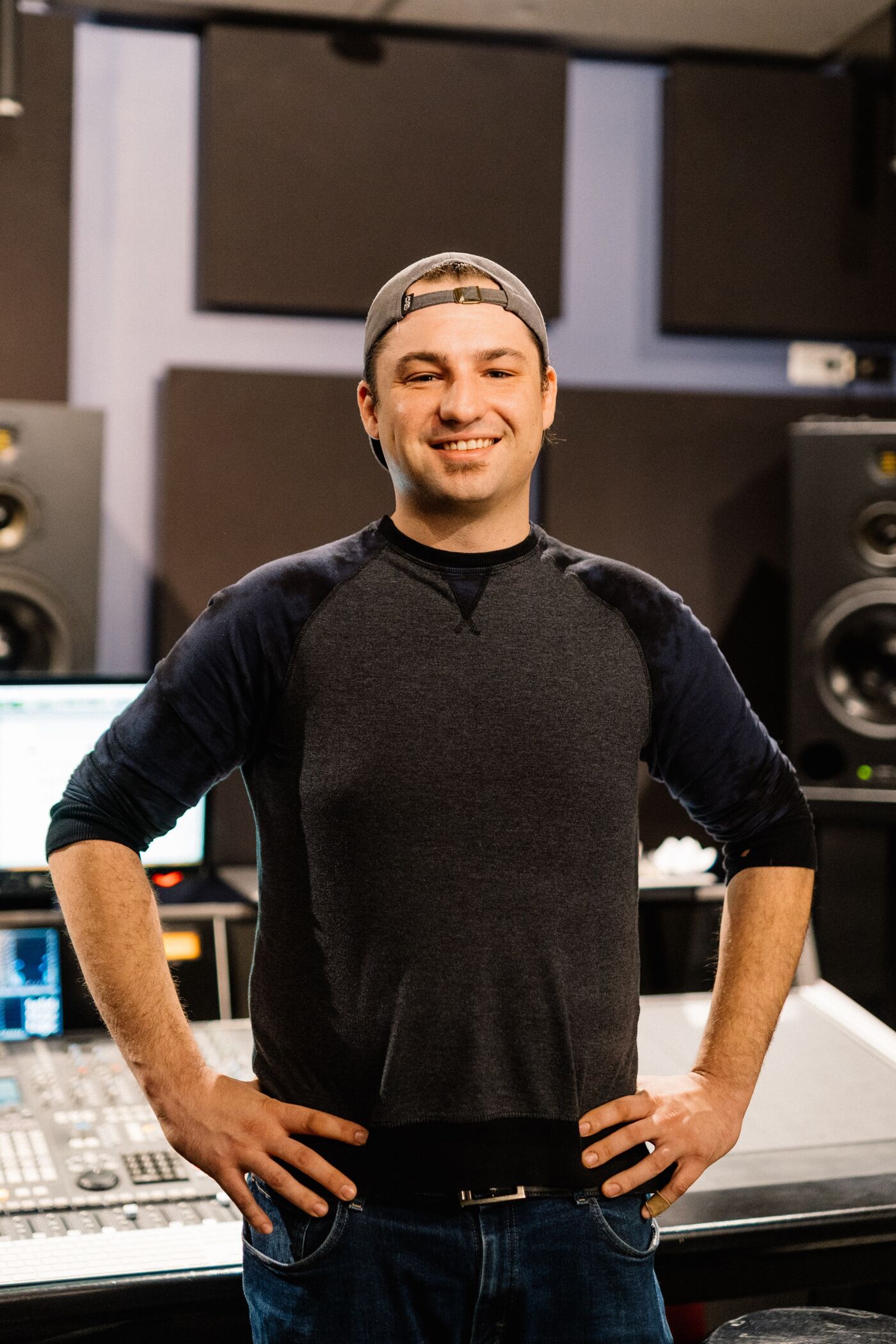A smiling audio engineer in casual attire stands confidently in a sound studio, hands on hips, ready to mix.