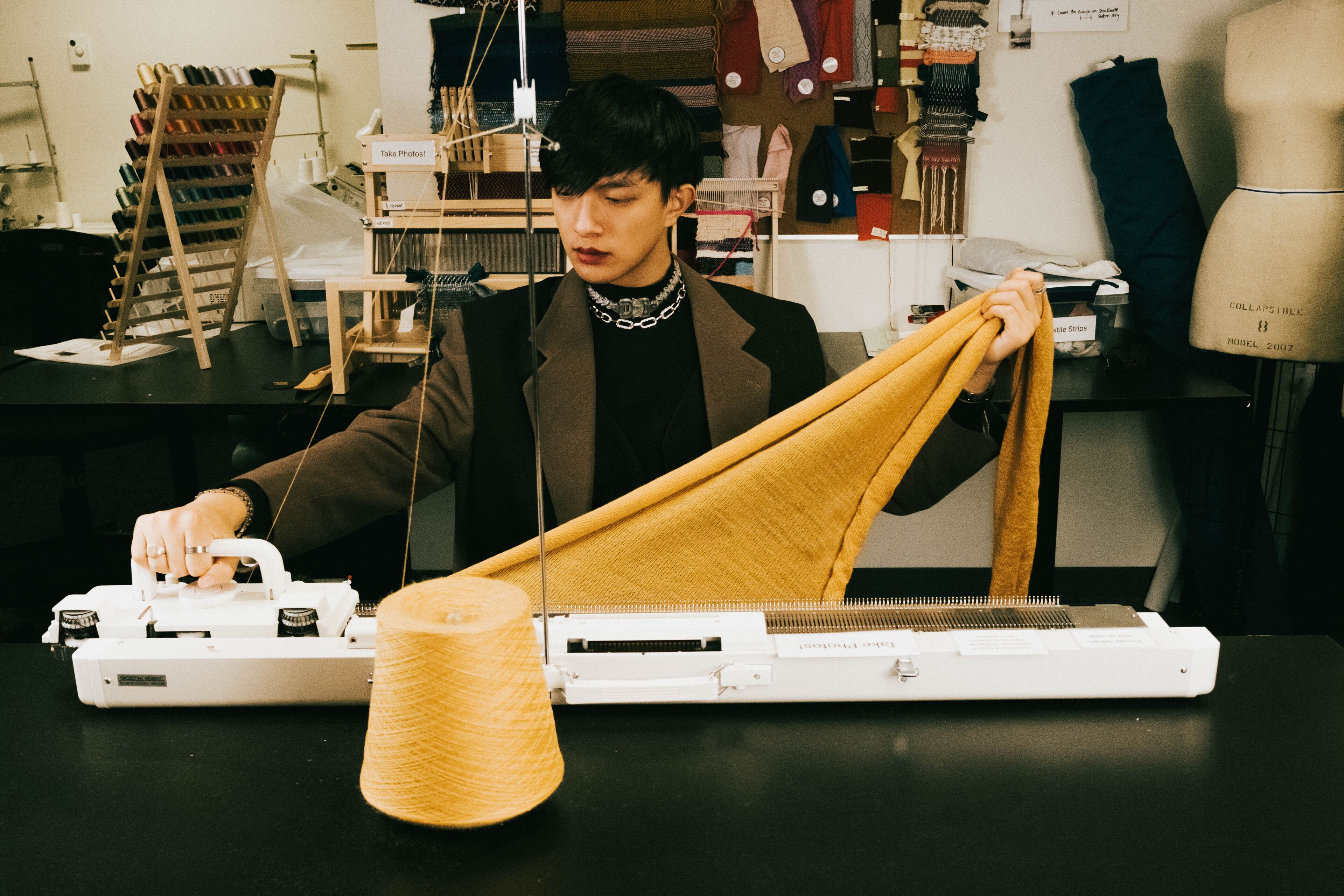 A focused designer operates a knitting machine, crafting a yellow textile piece, surrounded by a creative studio environment.