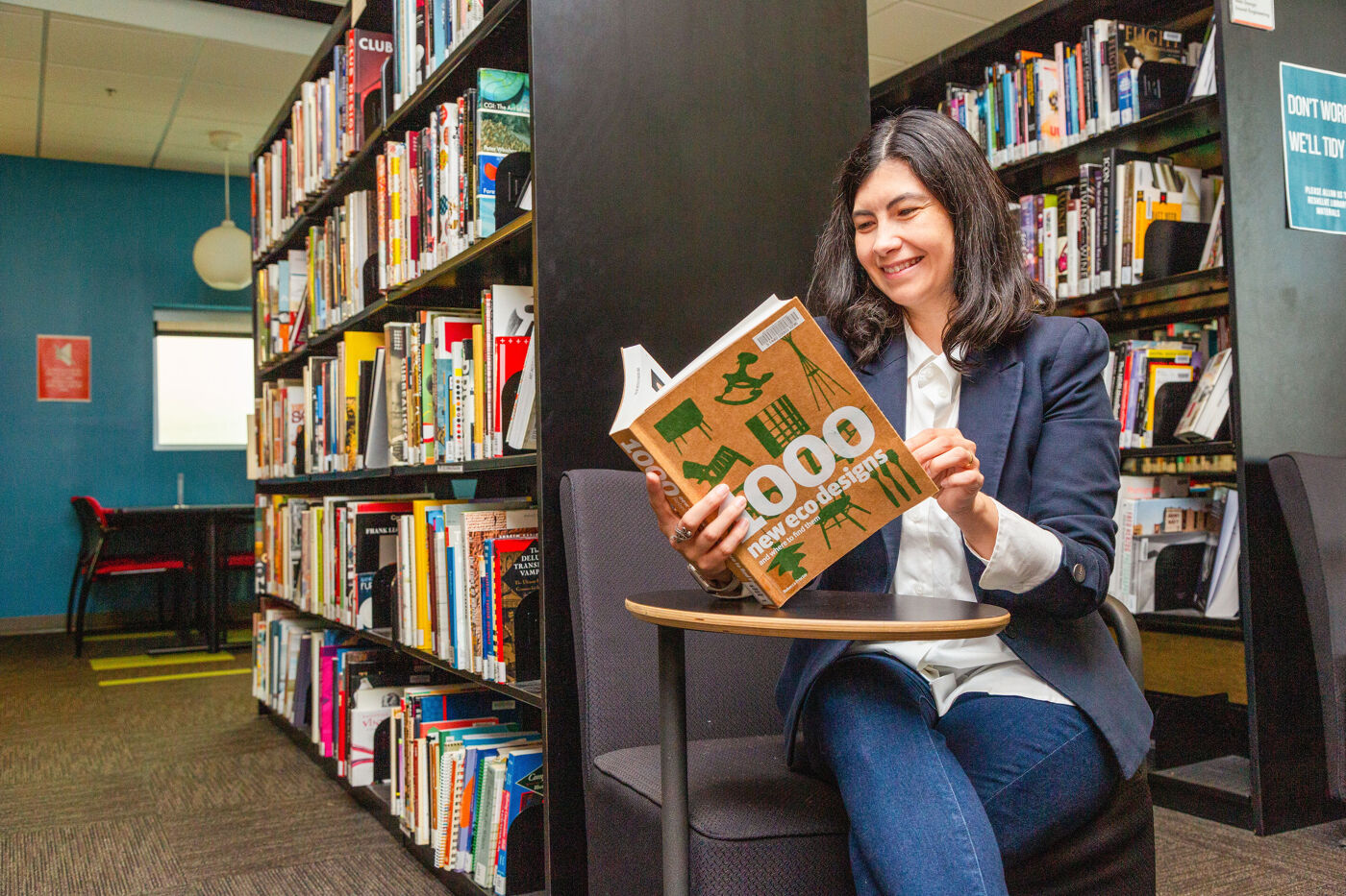 In the image, a woman is sitting at a table in a library, reading a book and smiling.