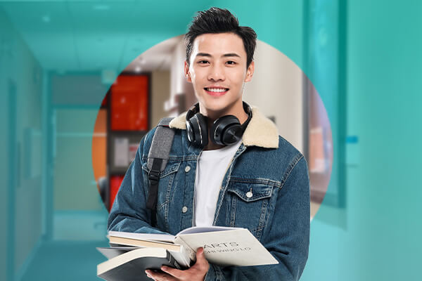A young man with headphones around his neck holding a book, standing in a school hallway.