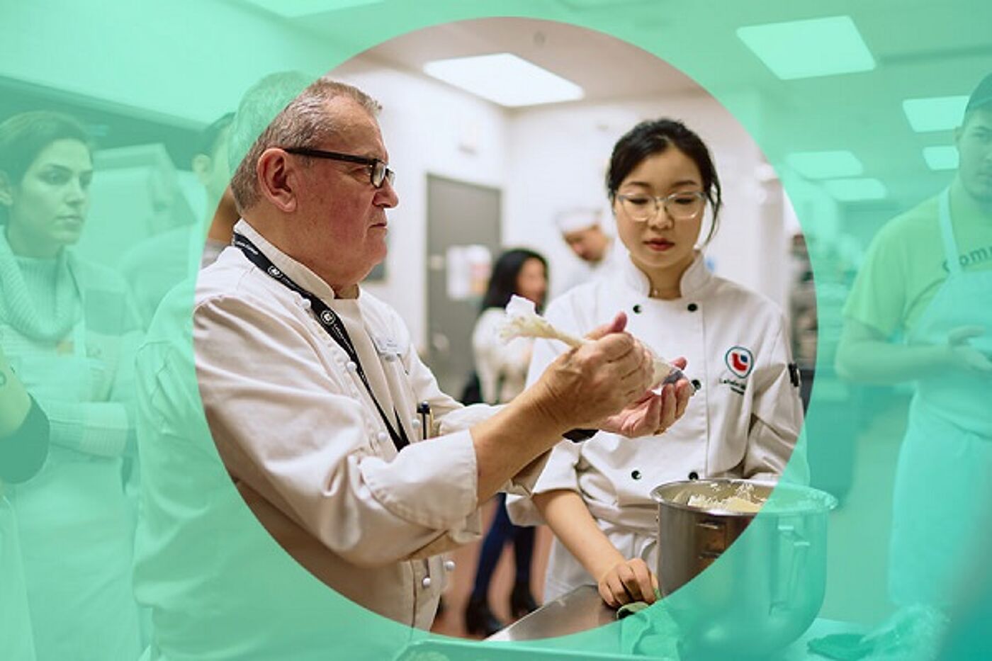 An experienced chef demonstrates a technique to a focused female student in a culinary class, surrounded by attentive learners.