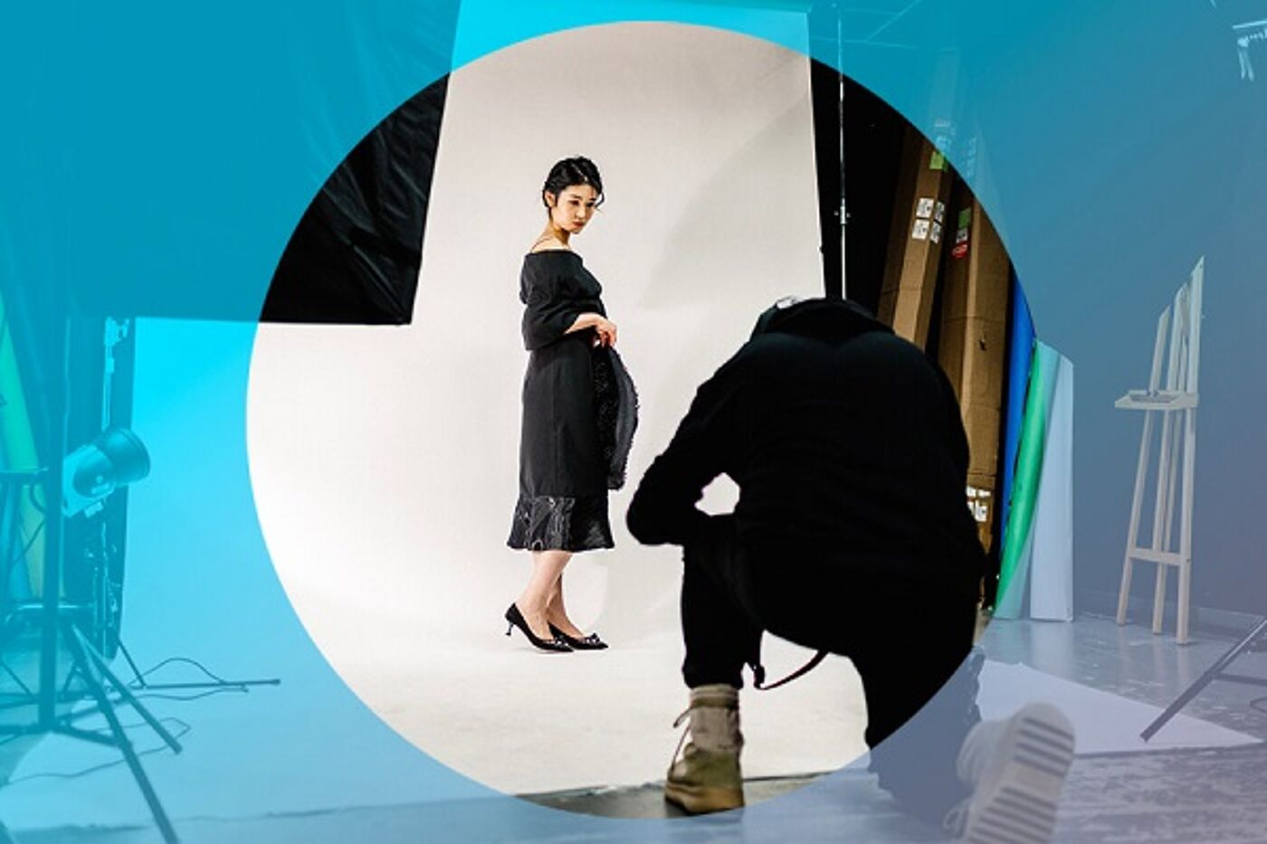 A model in a black dress poses for a photographer during a studio photoshoot, with lighting and equipment in the background.