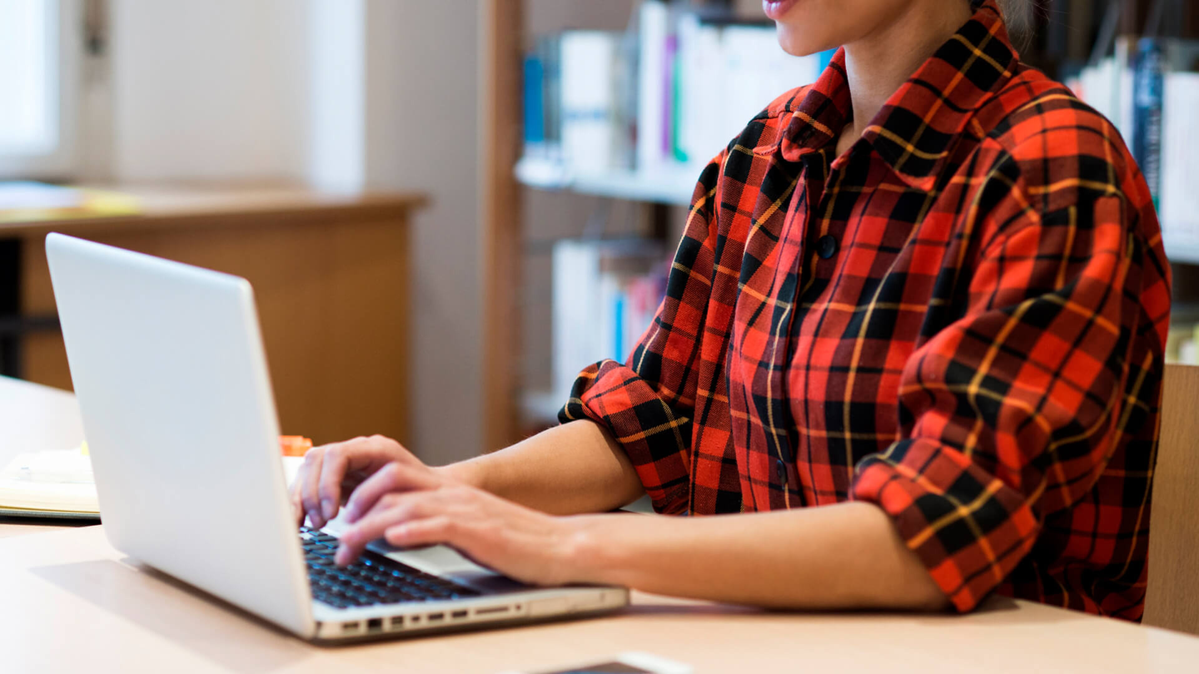 A focused individual in a red plaid shirt types on a laptop at a well-lit workspace, symbolizing productivity and the casual professional setting.