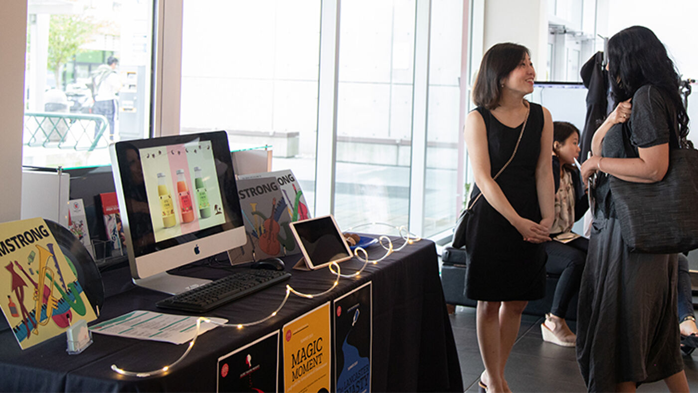 A table with a product showcase, including an iMac displaying colorful designs, promotional posters, and marketing materials, set up at a professional networking event. Two women engage in conversation nearby, highlighting a collaborative atmosphere.