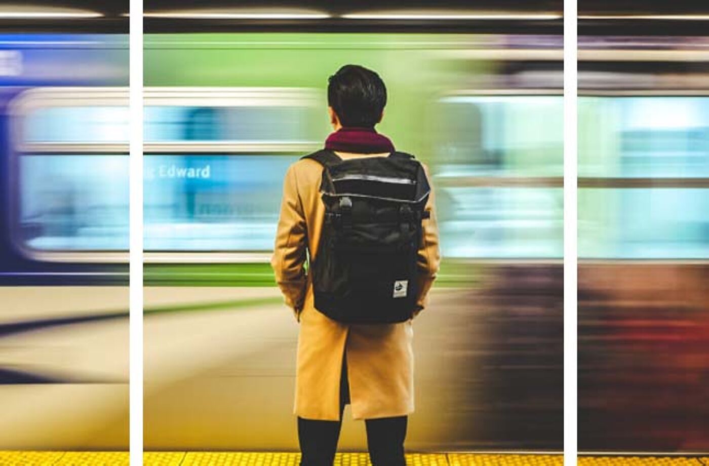 A man with a backpack and a beige coat stands on a subway platform as a train speeds past, creating a motion blur effect. The scene conveys urban transit and the daily rhythm of city life.