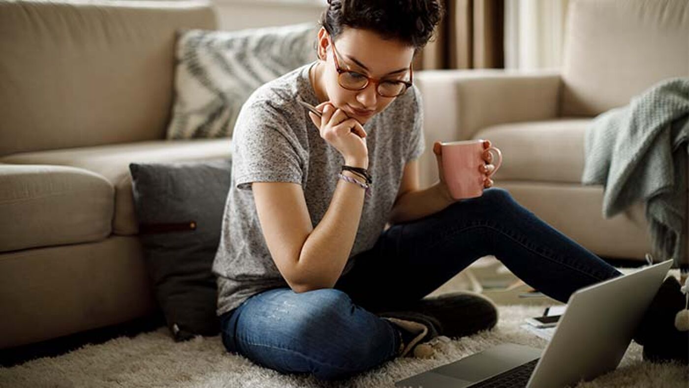 A focused woman sits on the floor, working intently on her laptop, with a mug in hand, suggesting a relaxed home environment.