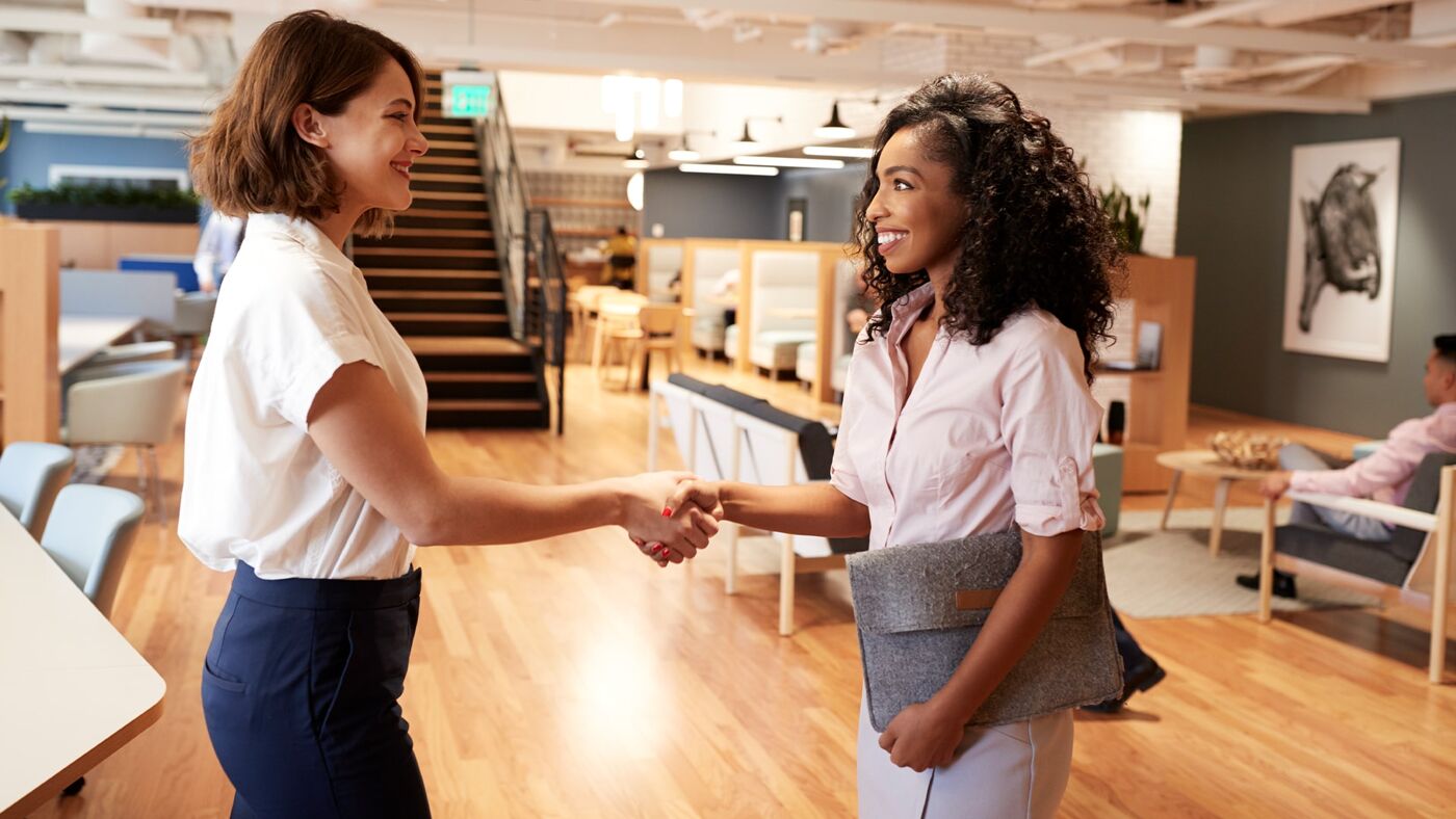 Two businesswomen smiling and shaking hands in a well-lit modern office space, indicating a positive interaction, with colleagues in the background.