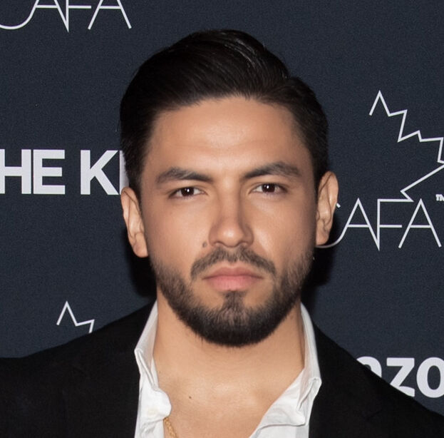 He wears a black suit with a white shirt, accessorized with a gold cross necklace, standing in front of a backdrop featuring logos such as Amazon, CAFA, and NARS.