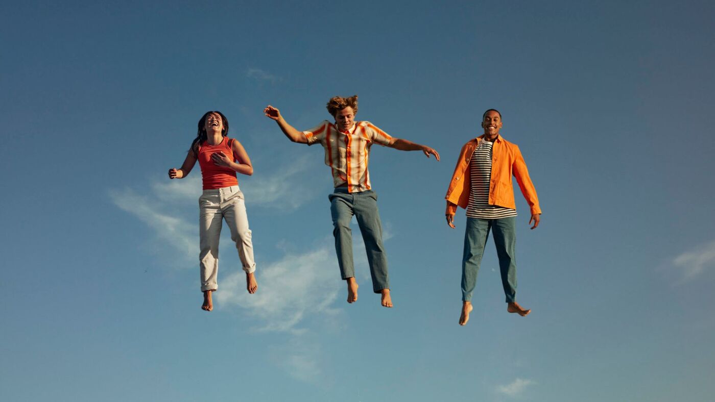 Three young adults joyfully leap against a clear blue sky.