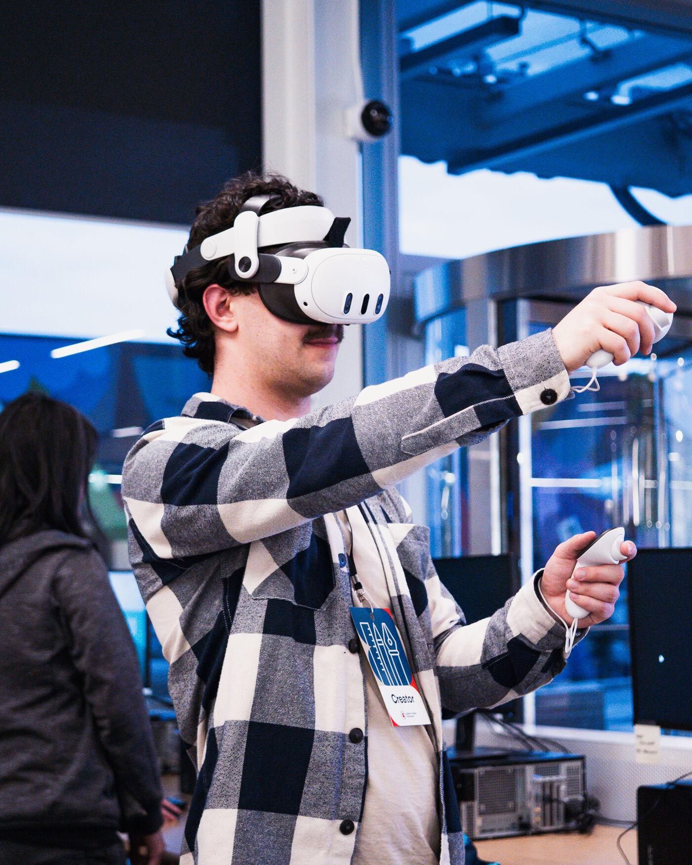 A man wearing a VR headset and holding controllers is immersed in a virtual reality experience. He is wearing a plaid shirt and a badge, and appears to be interacting with the virtual environment. A woman is standing to his left.