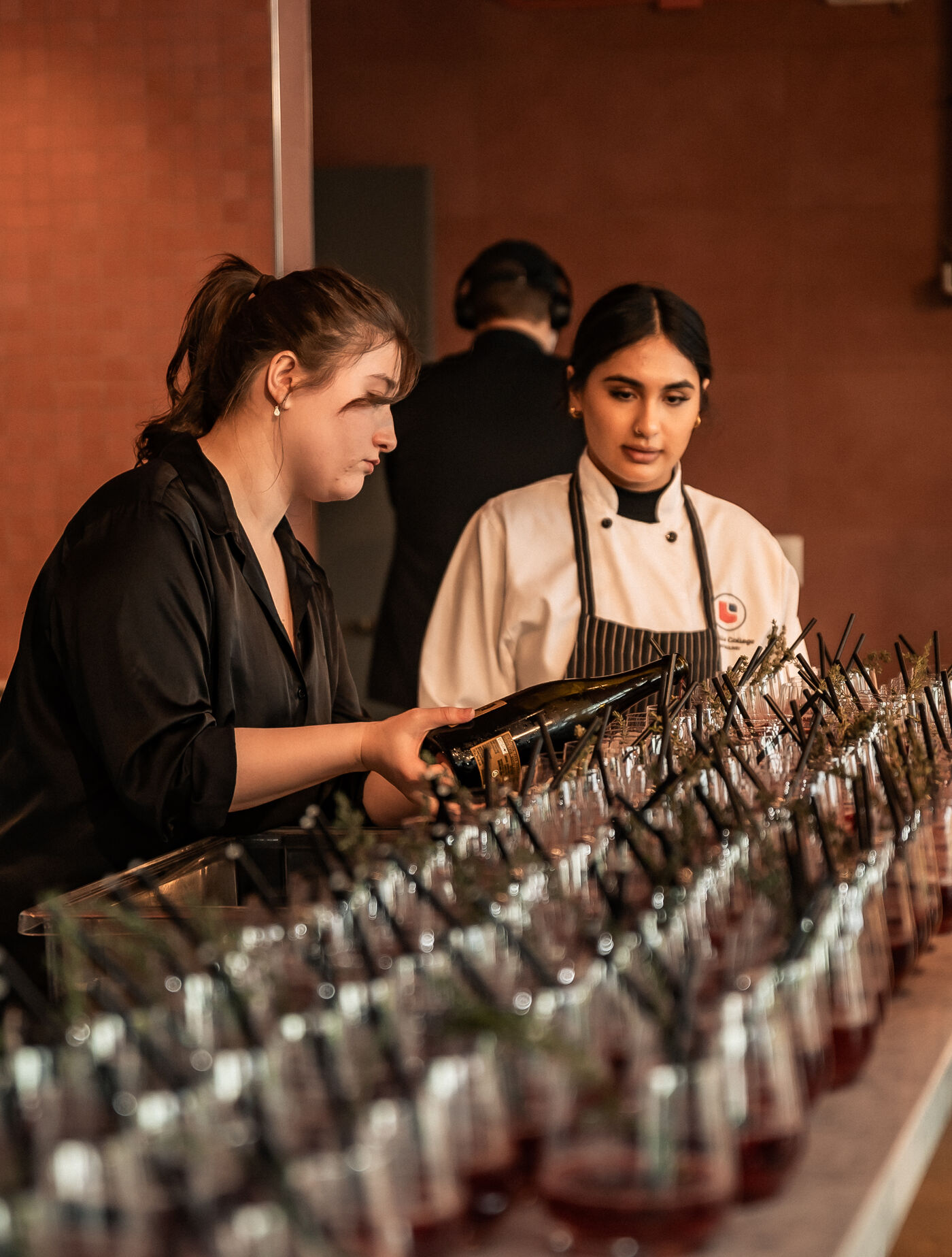 Two hospitality students, one in a chef's uniform, prepare for a beverage service by organizing glasses filled with red drinks garnished with herbs and straws. The setting is a professional service environment, reflecting attention to presentation and teamwork.