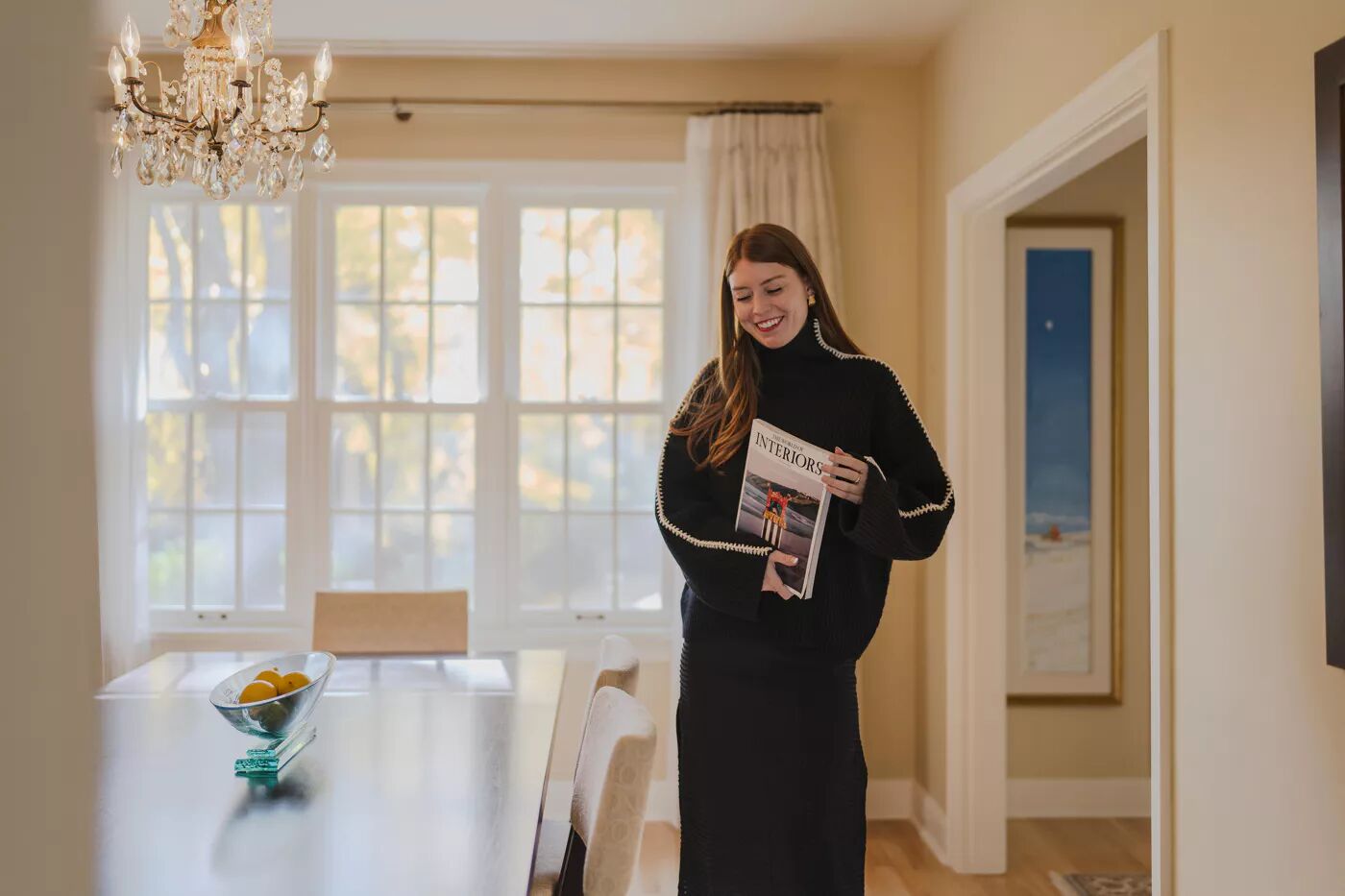 A woman in a black outfit stands in a dining room, holding a magazine. The room features a chandelier, a table with fruit, and a painting visible through a doorway. Natural light fills the space.