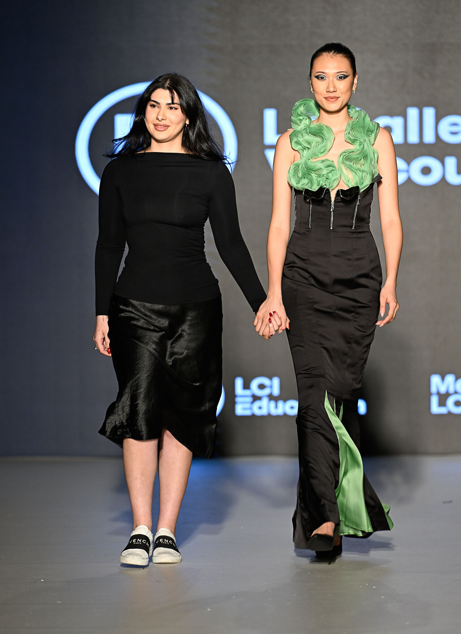 Two women are walking hand-in-hand on a runway. The woman on the left is wearing a black long-sleeved top and a black skirt, while the woman on the right is wearing a black dress with green accents.