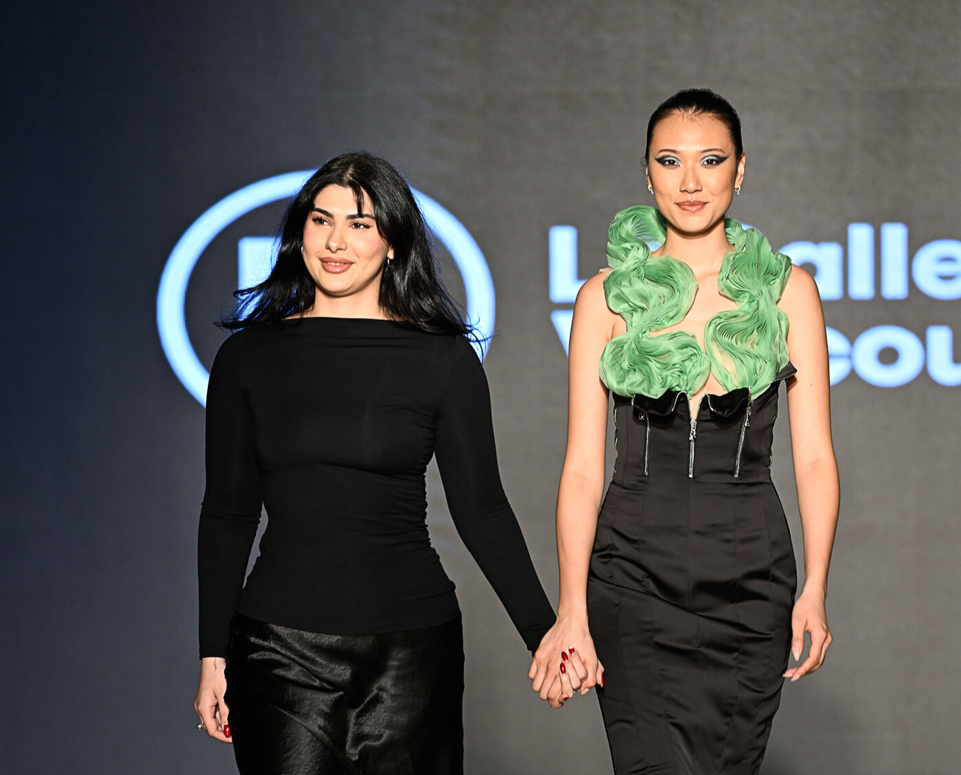 Two women are walking hand-in-hand on a runway. The woman on the left is wearing a black long-sleeved top and a black skirt, while the woman on the right is wearing a black dress with green accents.
