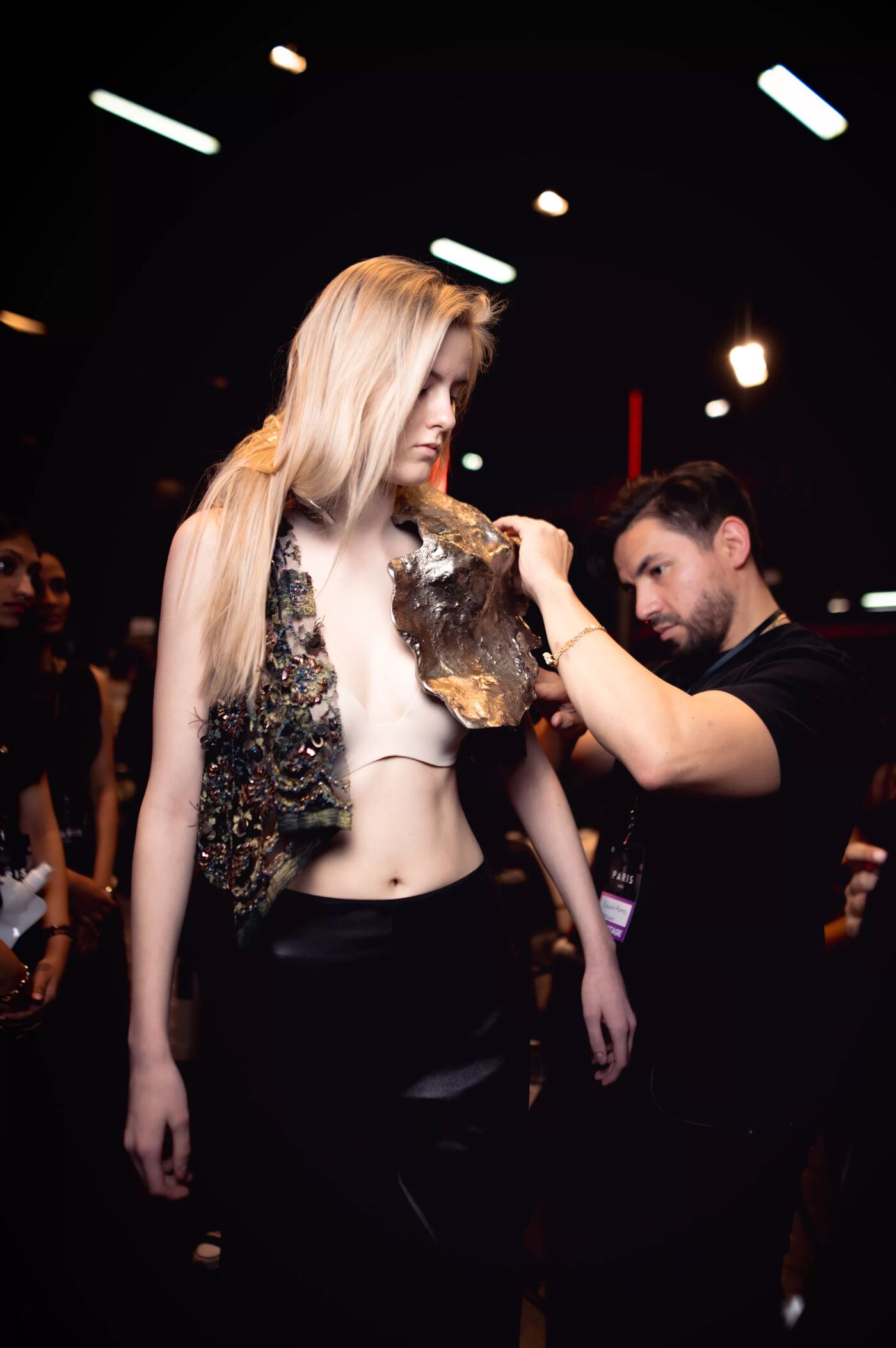 A model with long blonde hair is being adjusted by a man in a black shirt. She is wearing a unique, textured vest and black pants, possibly backstage at a fashion show. The lighting is dim, and other people are visible in the background.