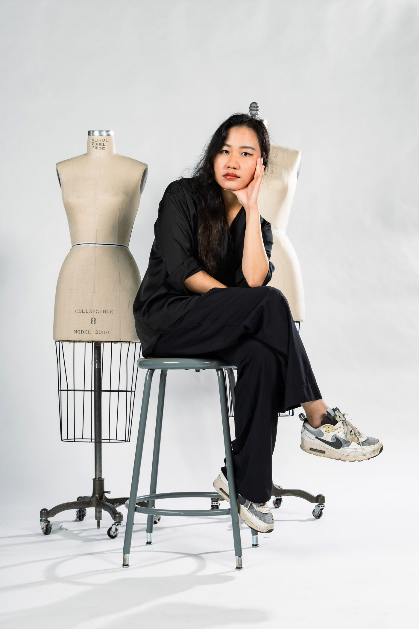 A woman with long dark hair is sitting on a stool, wearing a black outfit and sneakers. Behind her are two dress forms on stands. The background is a plain white color.