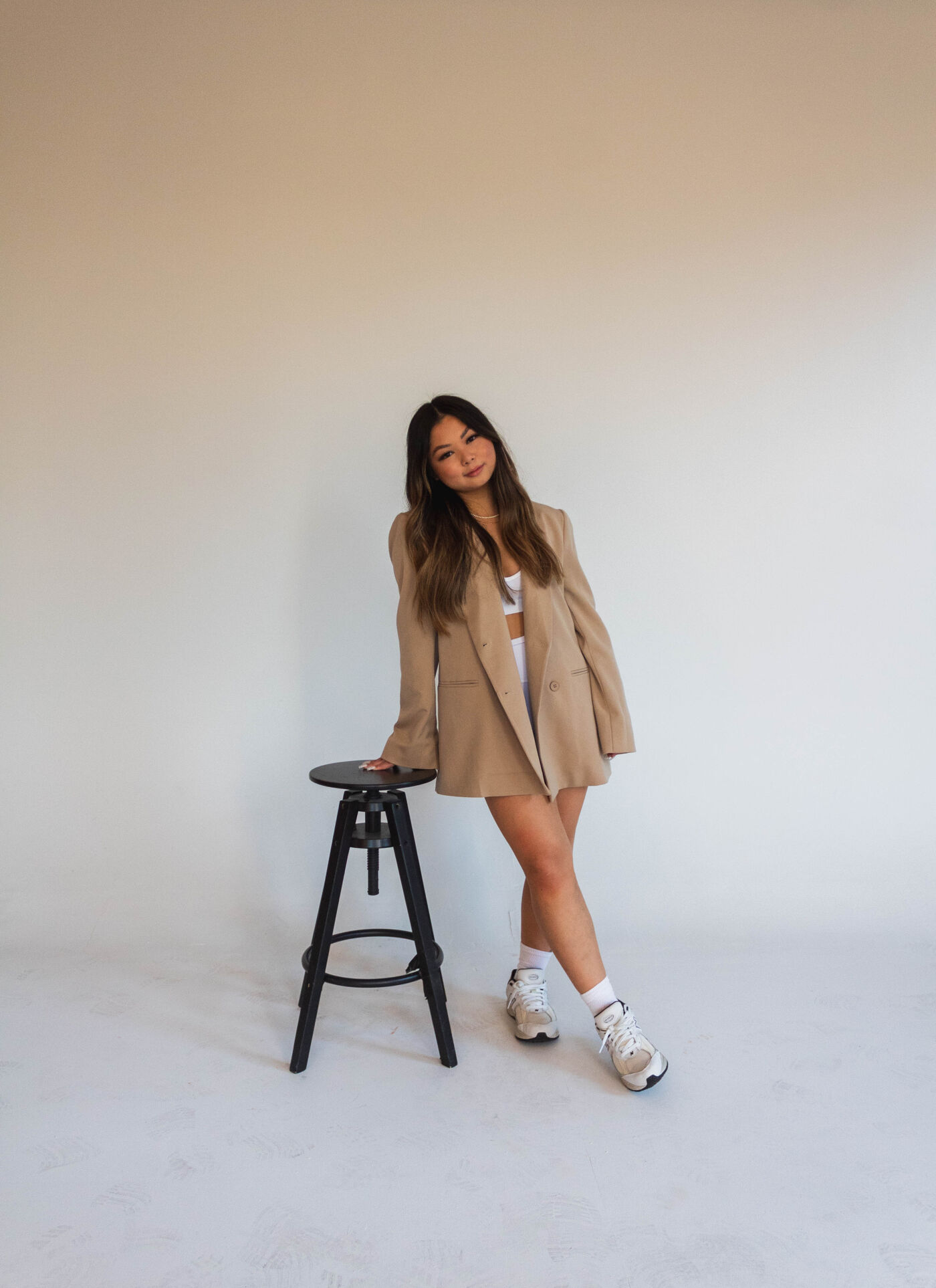 A young woman with long brown hair is posing next to a black stool. She is wearing a tan blazer, white socks, and white sneakers. The background is a plain white wall, creating a simple and clean aesthetic.