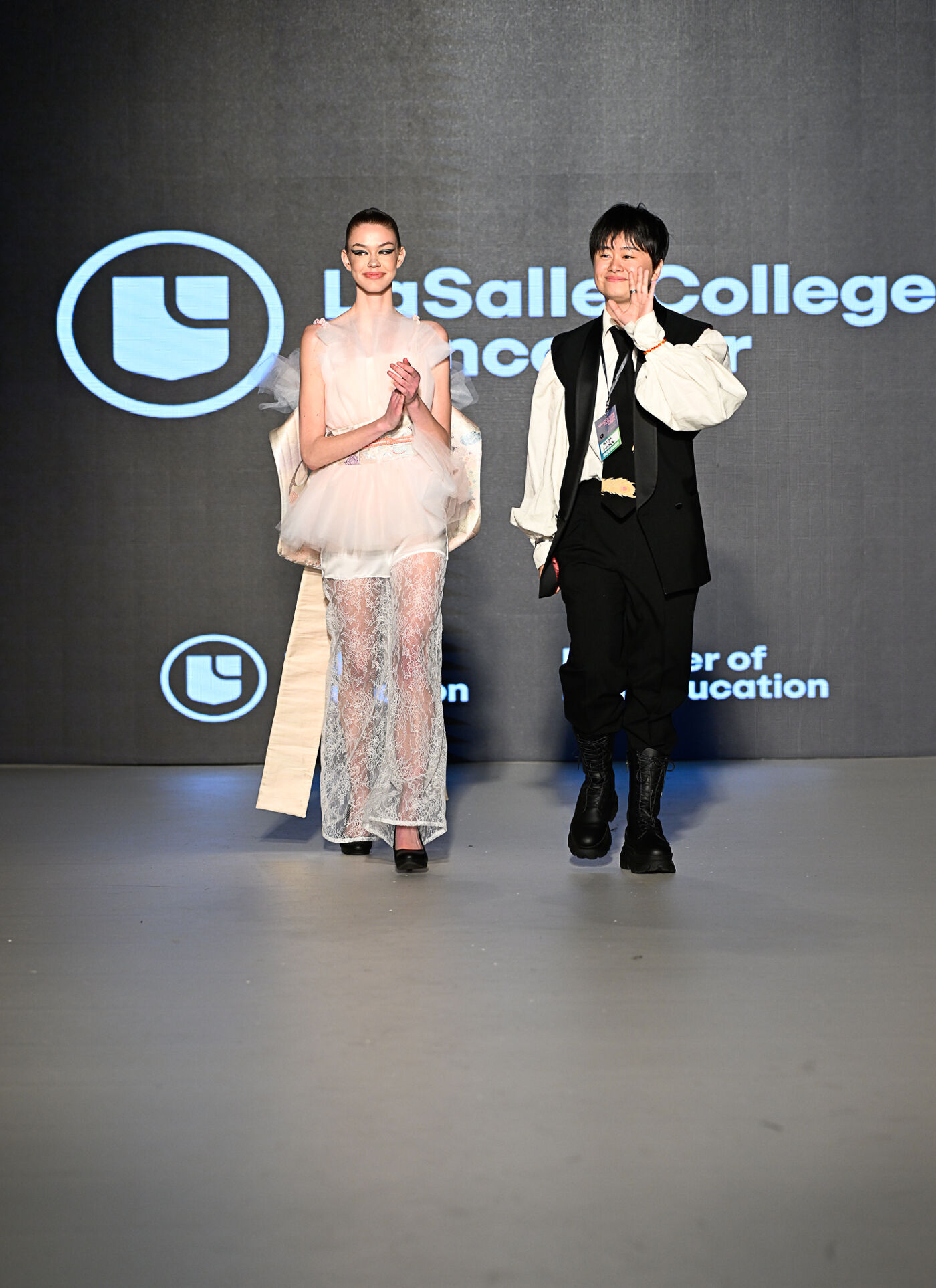 Two models are walking on a runway. The model on the left is wearing a light pink dress with a tulle skirt, and the model on the right is wearing a black vest and pants with a white shirt.