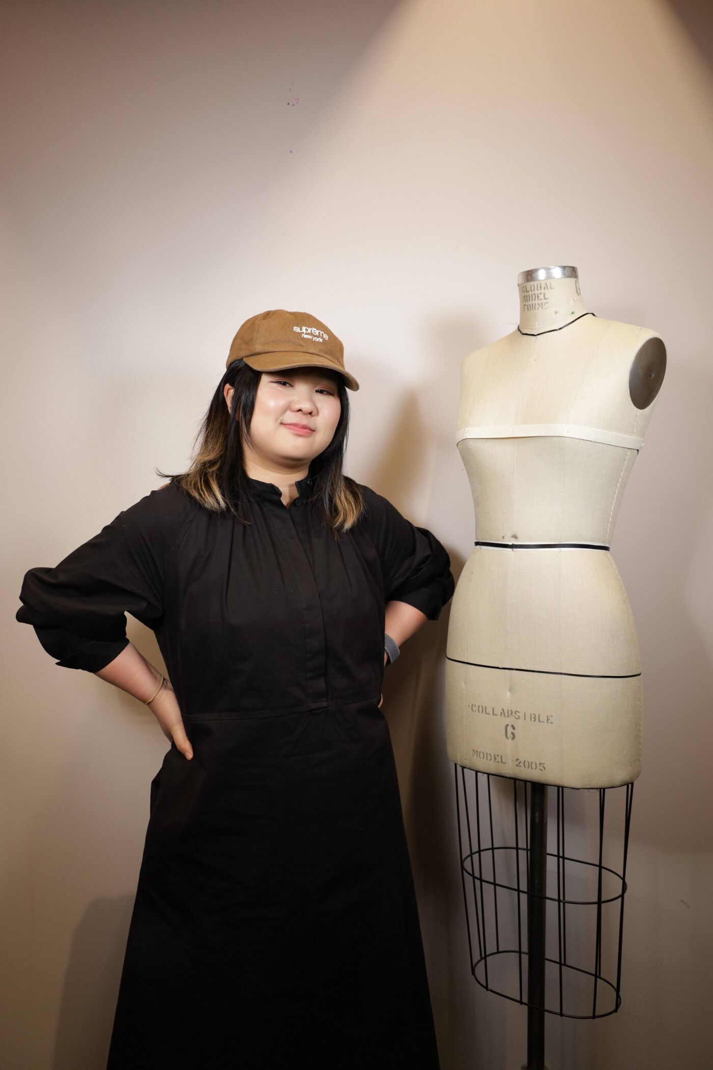 A woman wearing a black dress and a brown cap stands next to a mannequin. The mannequin is a dress form with a wire cage base. The woman has her hand on her hip and is smiling slightly.