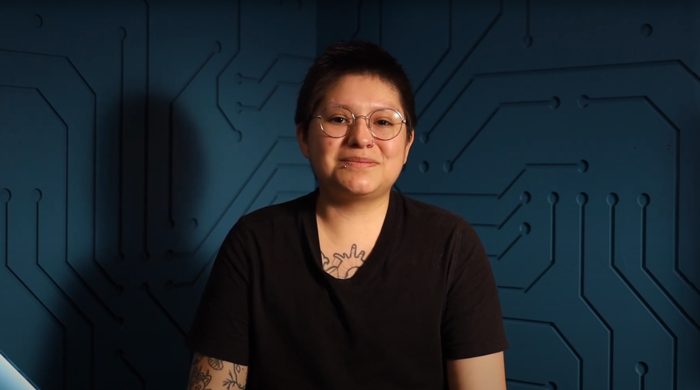 The image shows a person with short dark hair, glasses, and a black t-shirt, smiling slightly at the camera. They have tattoos on their chest and arm. The background is a blue wall with a circuit board pattern.