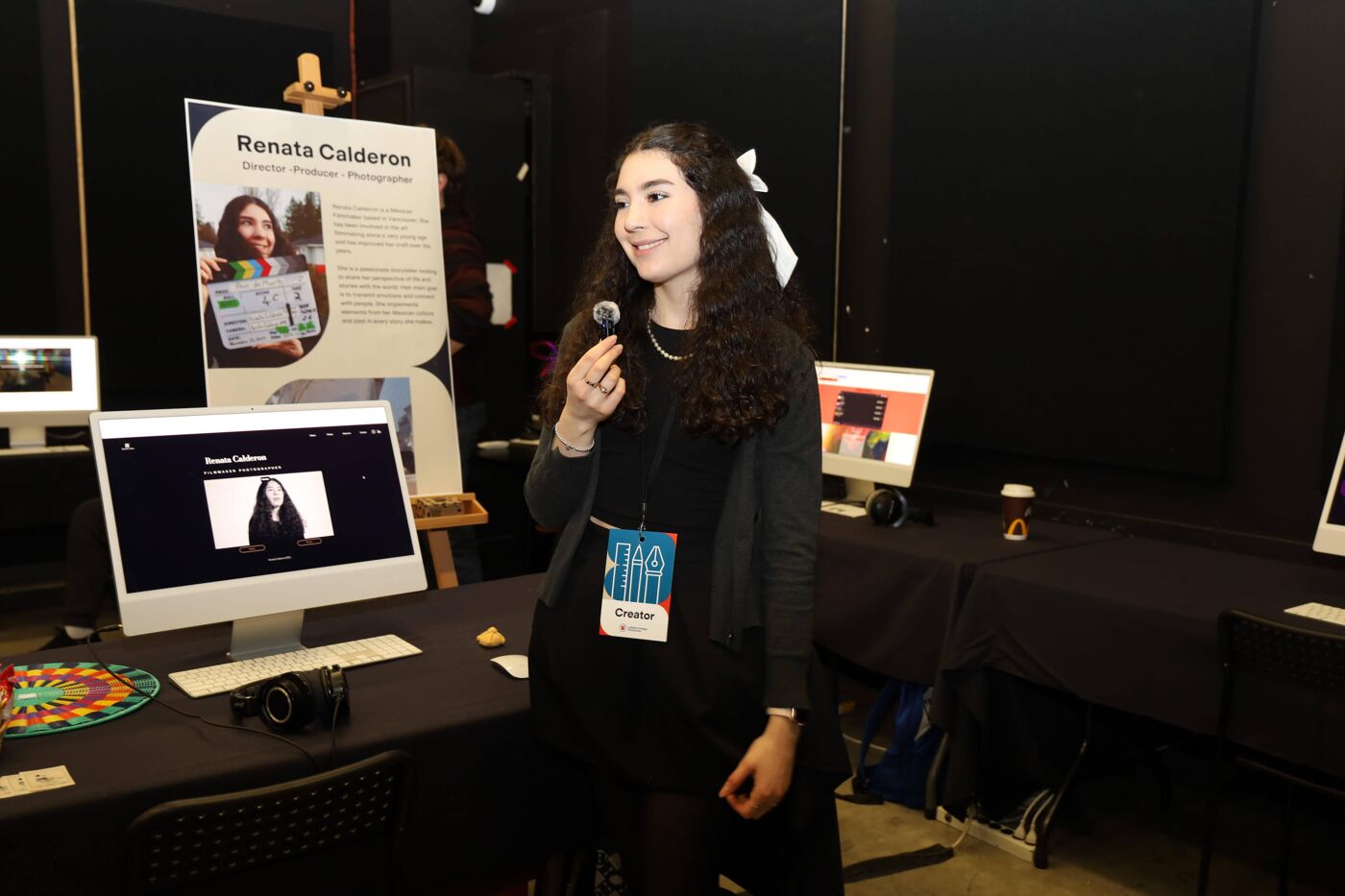 A woman with long dark hair is standing at a booth, holding a microphone and smiling. Behind her is a poster with a photo of a woman and text. There is also a computer monitor on the table in front of her.