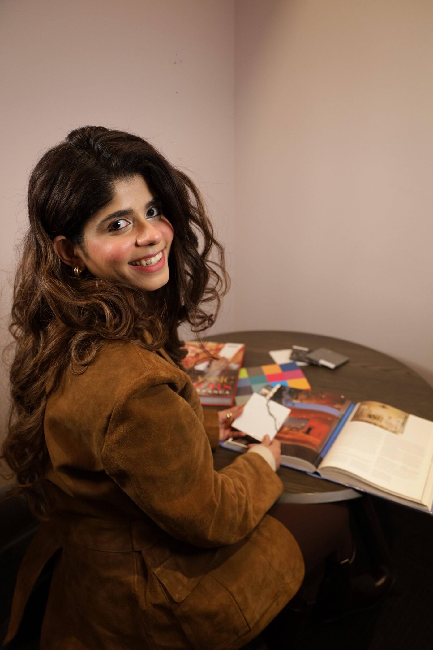 A woman with long, wavy brown hair smiles at the camera while sitting at a round table. She is wearing a brown jacket and surrounded by books, cards, and other items, suggesting she may be studying or working on a project.