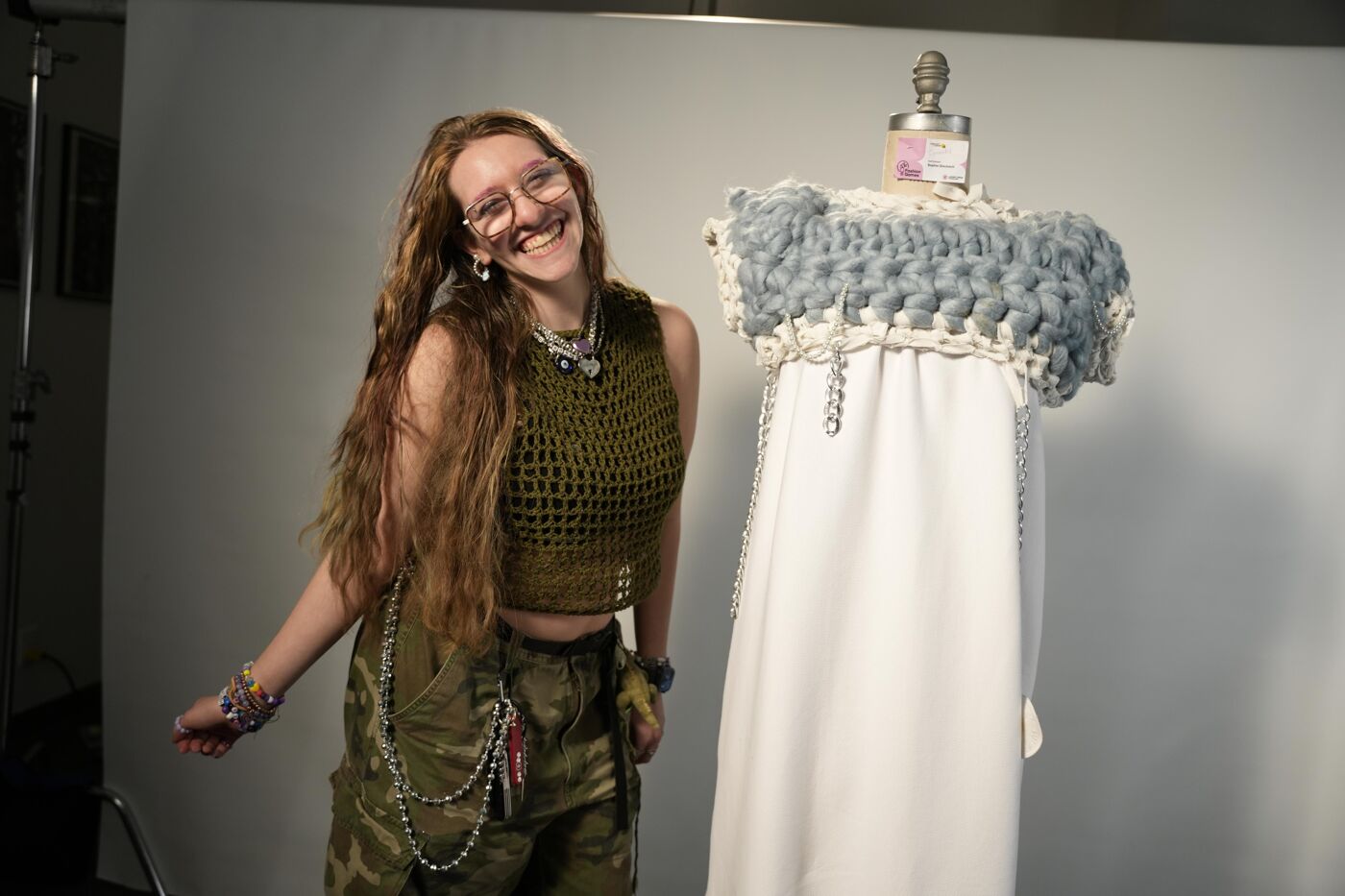 A smiling woman with long hair stands next to a white dress form. She is wearing a green crocheted top, camouflage pants, and glasses. The dress form is adorned with a blue and white crocheted top.