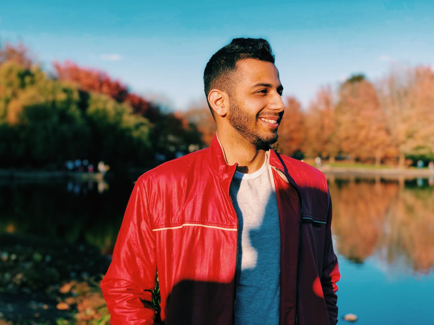 A man in a red jacket smiles as he stands in front of a lake surrounded by trees with autumn foliage. The sky is blue and the sun is shining, creating a pleasant outdoor scene.