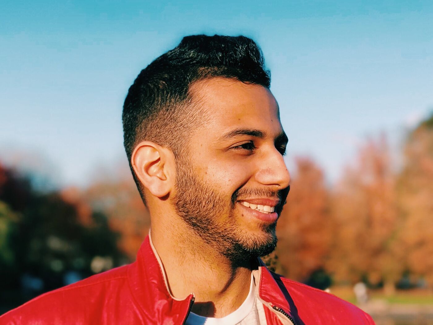 A man in a red jacket smiles as he stands in front of a lake surrounded by trees with autumn foliage. The sky is blue and the sun is shining, creating a pleasant outdoor scene.