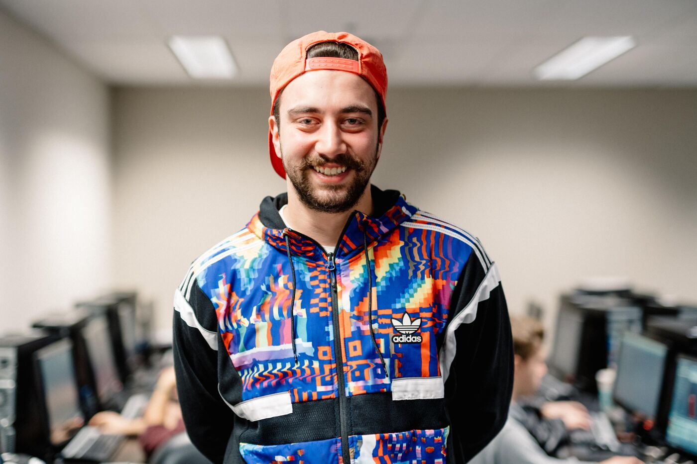 A man with a beard and an orange cap is smiling at the camera. He is wearing a colorful Adidas jacket with black and white accents. The background shows a room with computers and other people. 