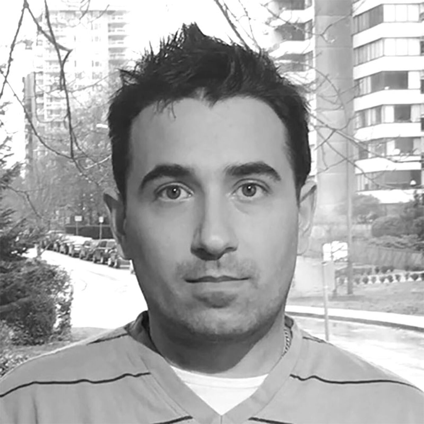 Close-up black and white shot of a man with short, dark hair, wearing a horizontally striped shirt, standing outdoors with a blurred city background.