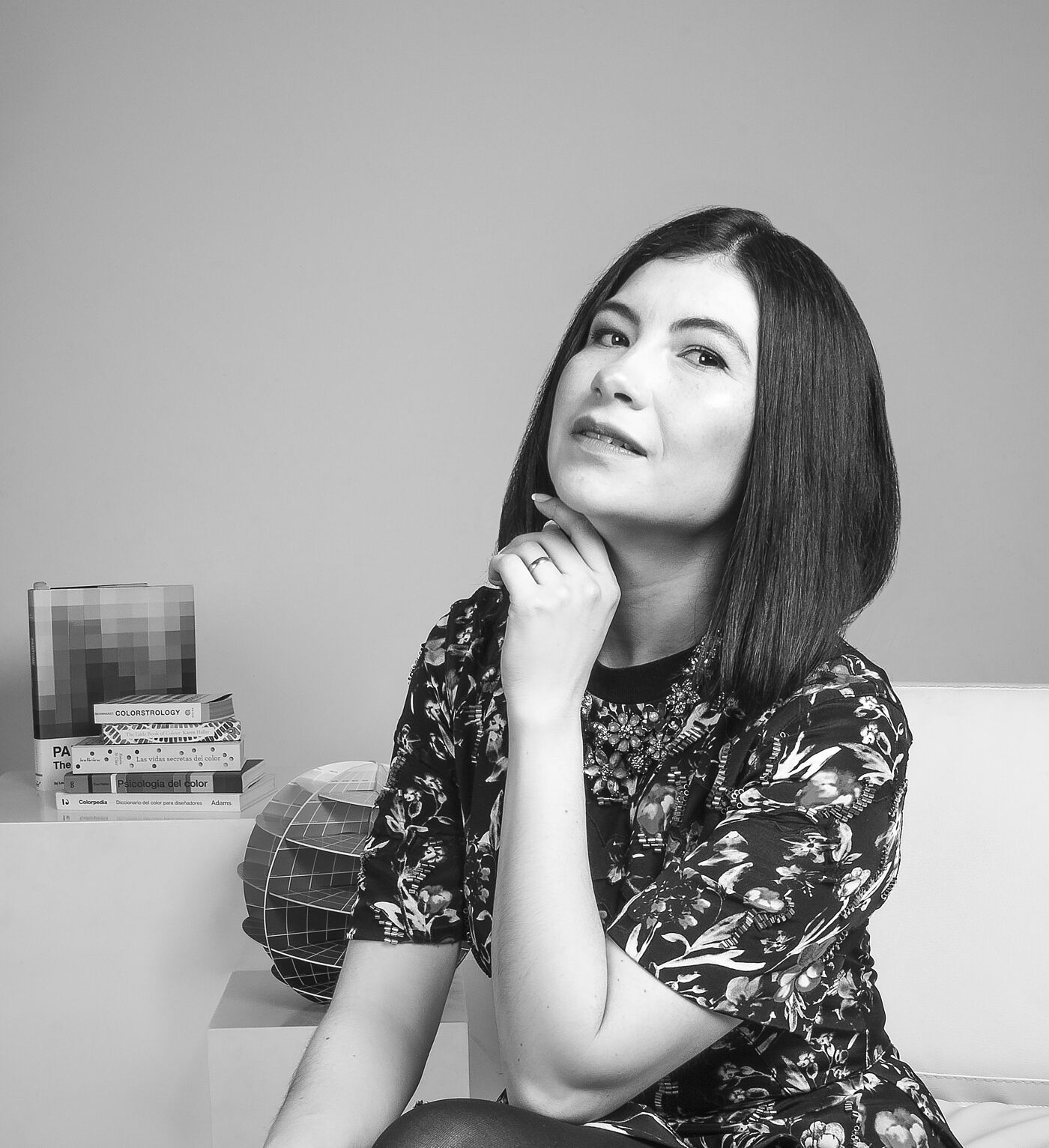 A black and white, medium shot of a woman in a floral dress and black tights, sitting on a white couch, thoughtfully touching her chin.