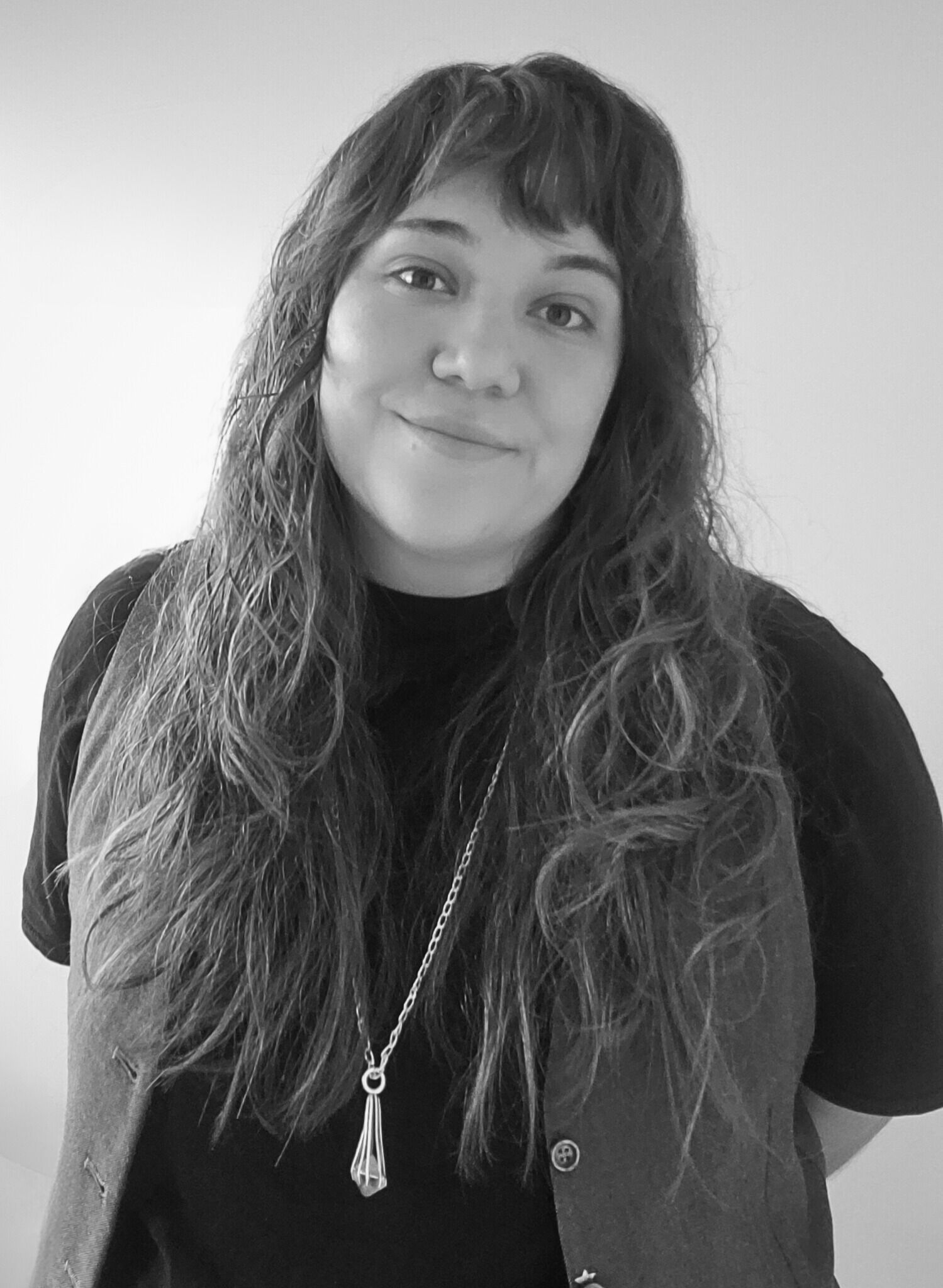 Close-up black and white shot of a woman with long, wavy hair wearing a black shirt and vest, and a silver necklace.