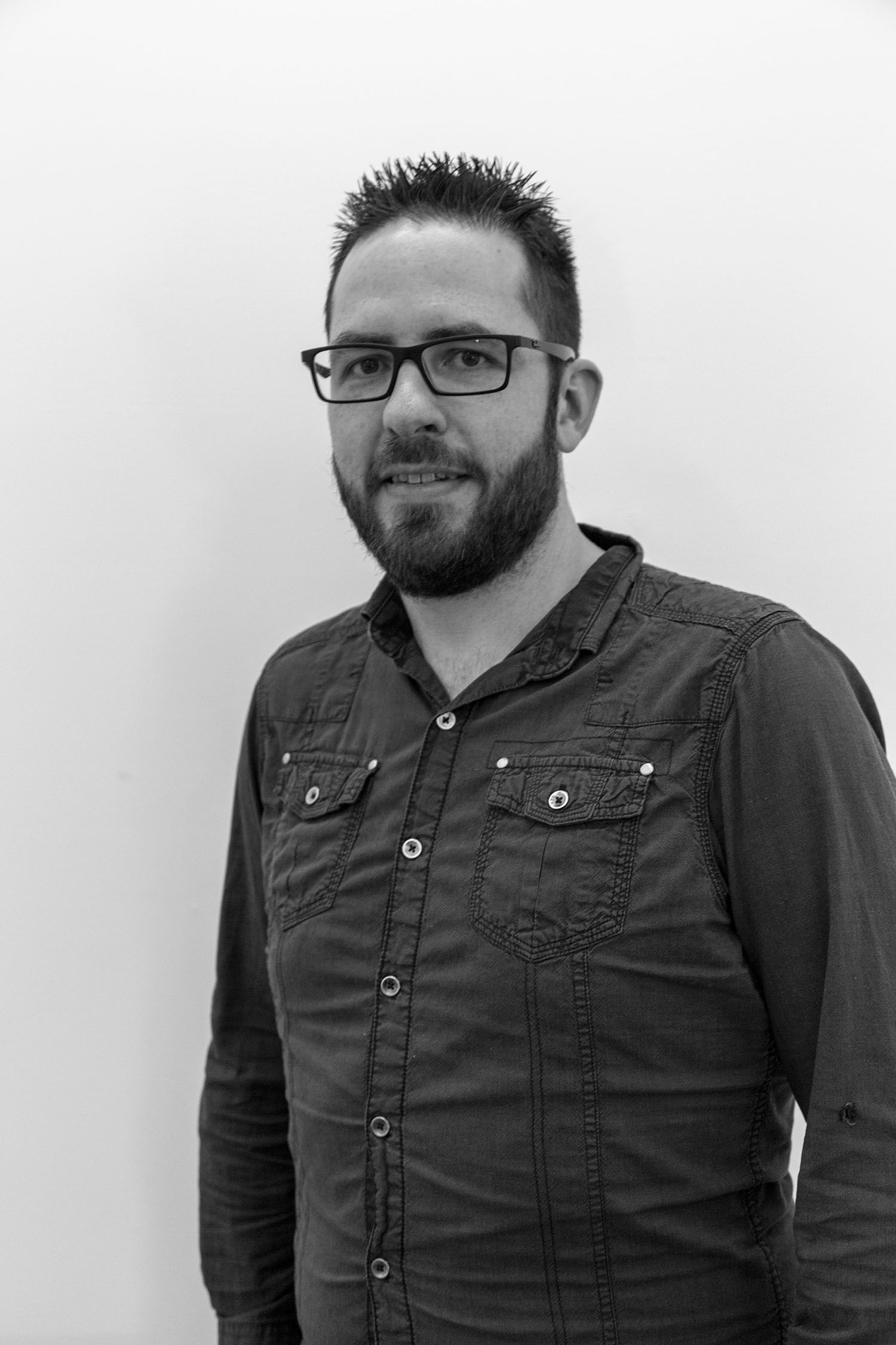 Close-up black and white shot of a man with glasses and a beard, wearing a dark button-down shirt.