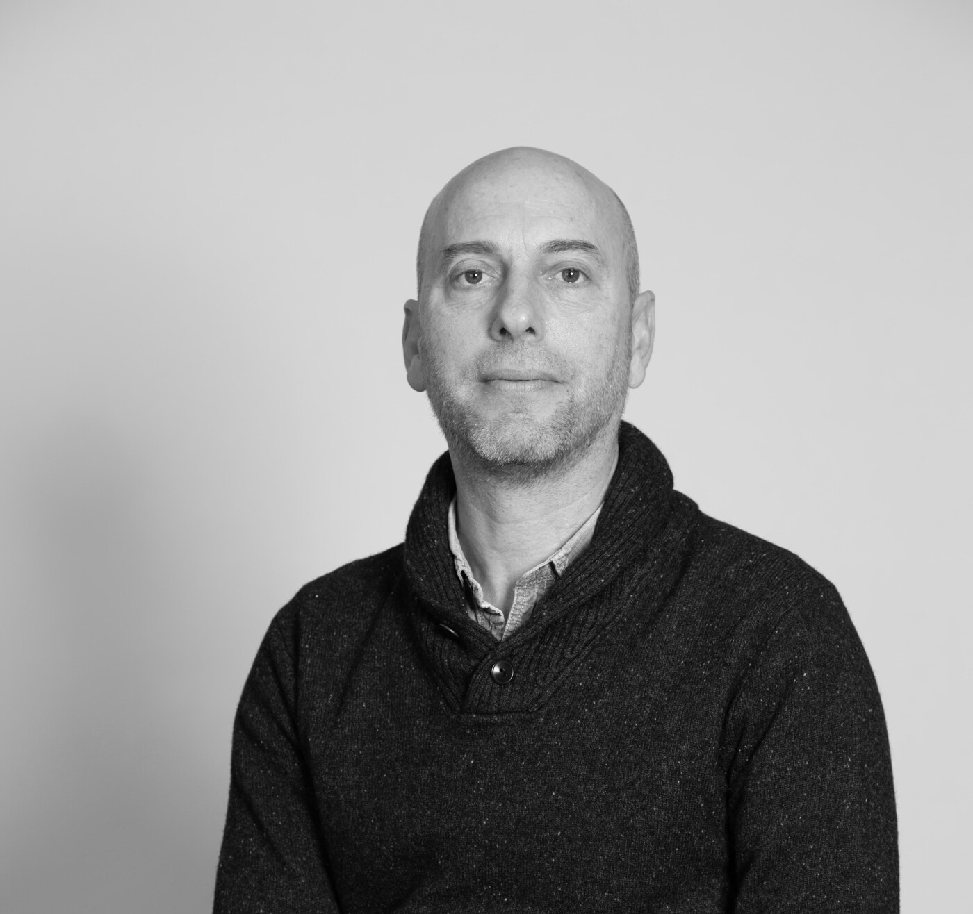 A black and white, eye-level, medium shot of a bald man with a serious expression sitting and wearing a dark-colored collared sweater.