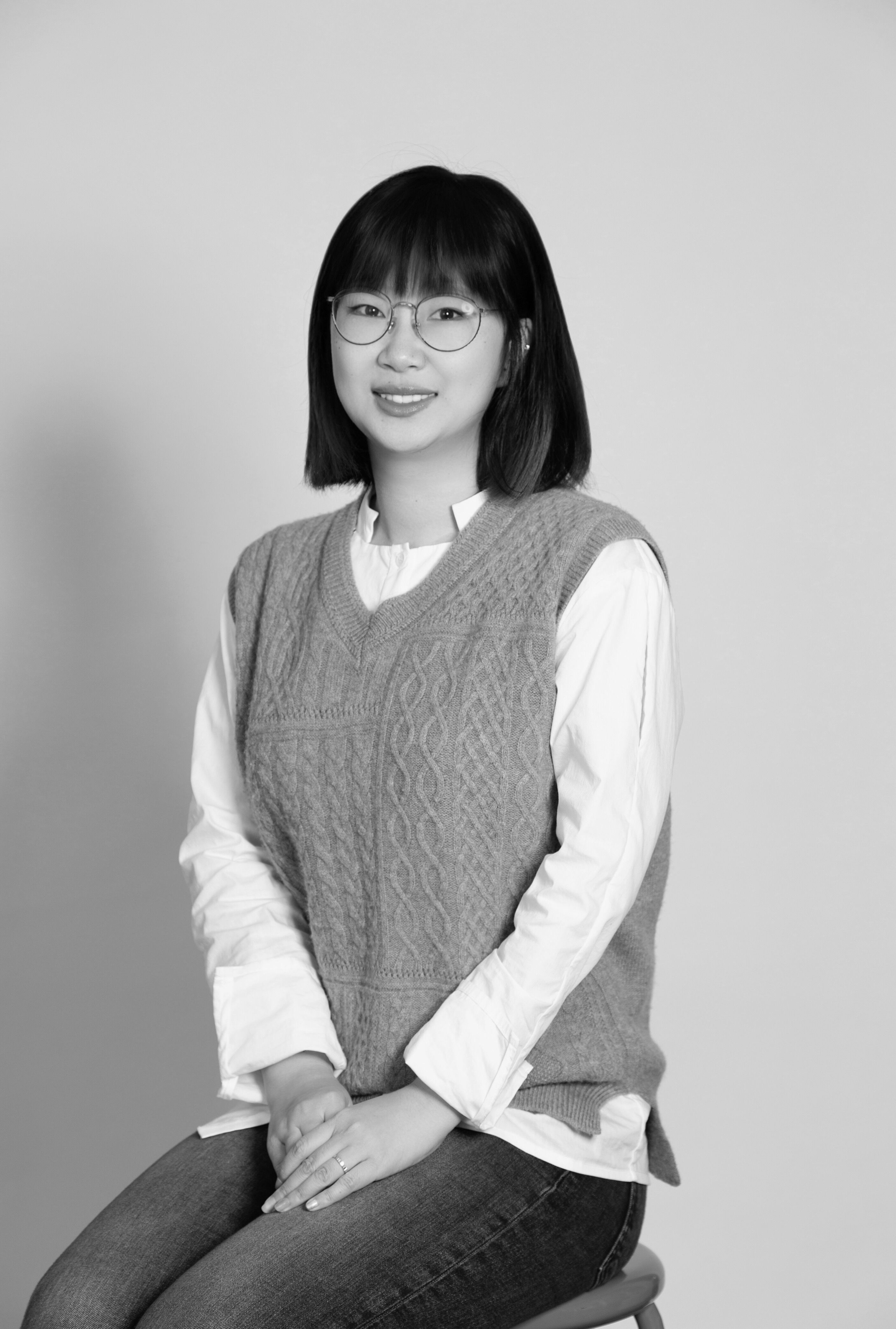 A black and white, eye-level, medium indoor shot depicts a young woman with short, straight, dark hair, glasses, and a light-colored sweater-vest over a white long-sleeved shirt, sitting on a stool against a light-yellow background.