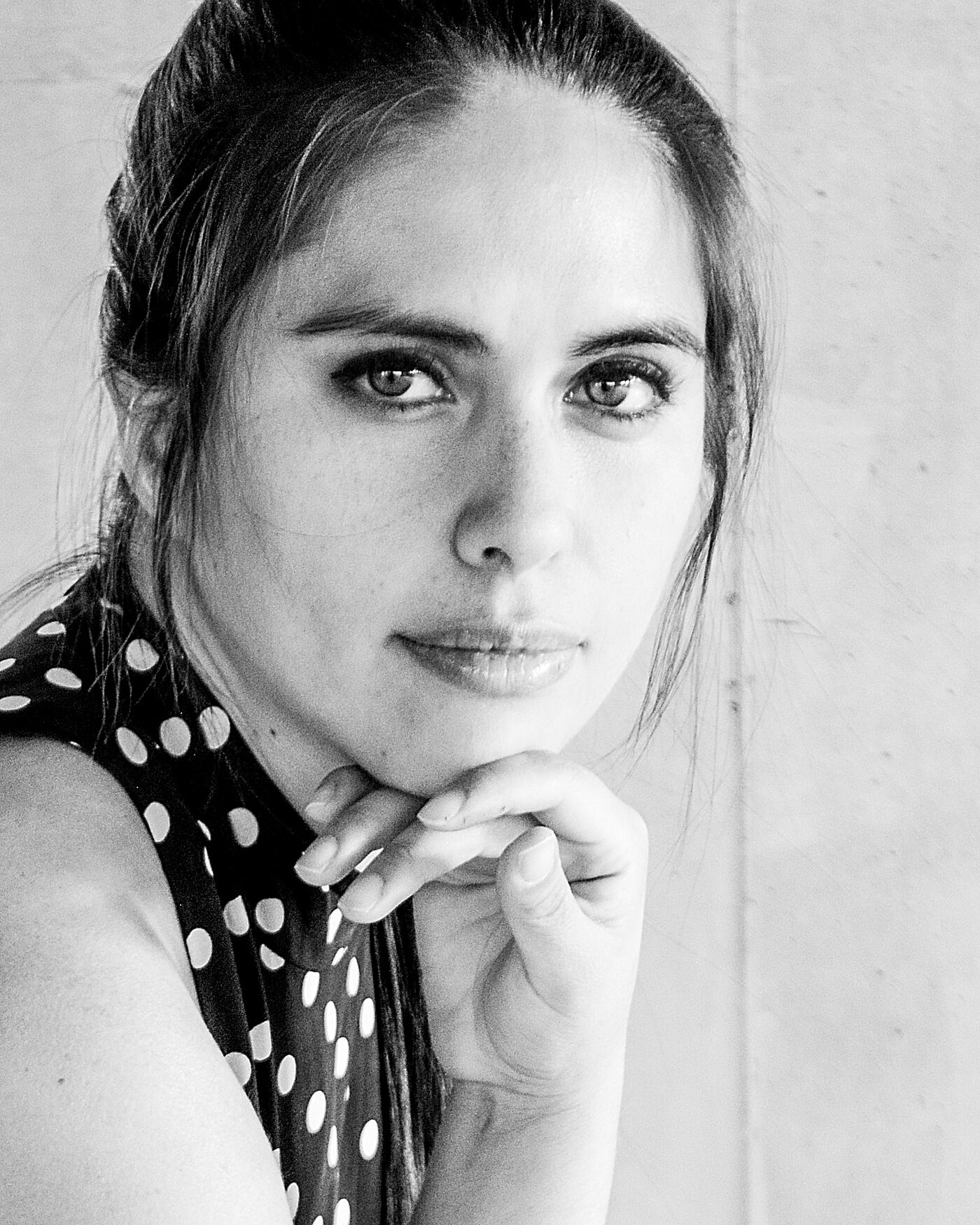 Close-up black and white shot of a woman with dark hair pulled back, wearing a polka dot top, her hand resting on her chin.