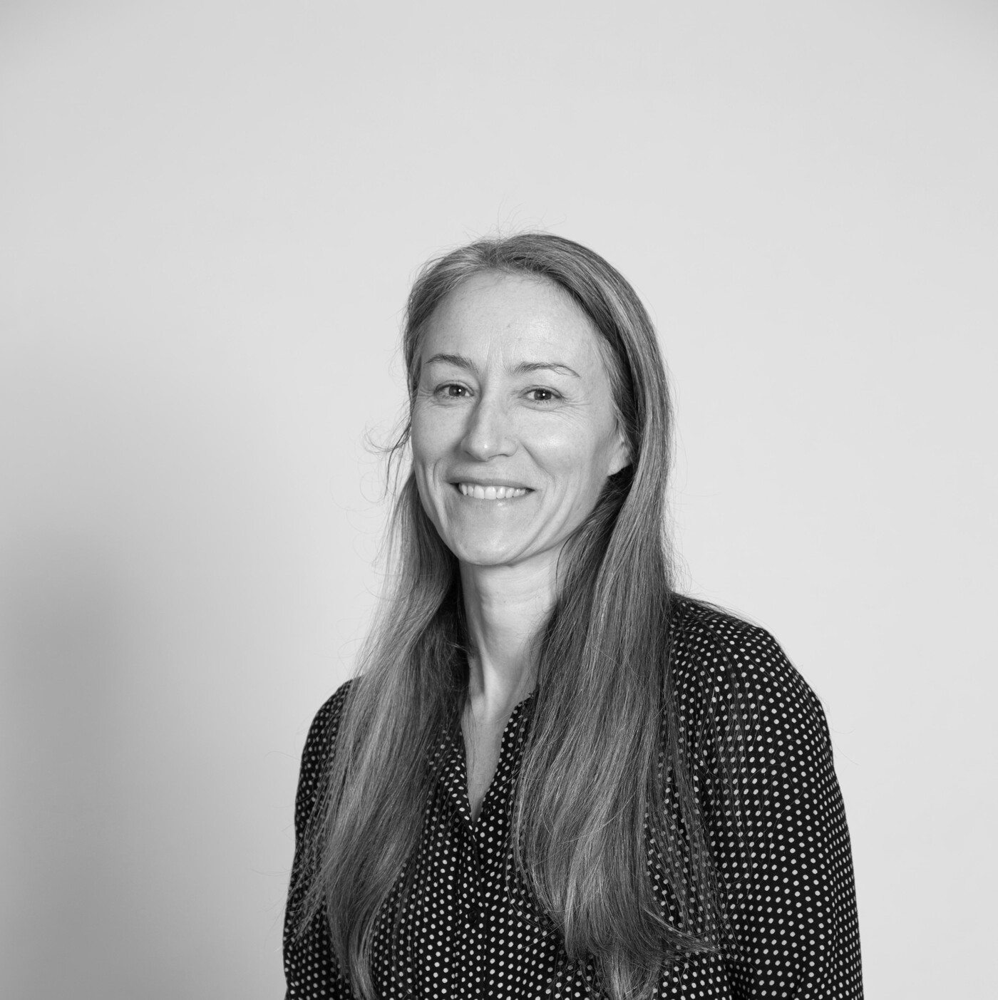 A black and white, eye-level, medium shot of a woman with long hair wearing a polka dot shirt, sitting and smiling.