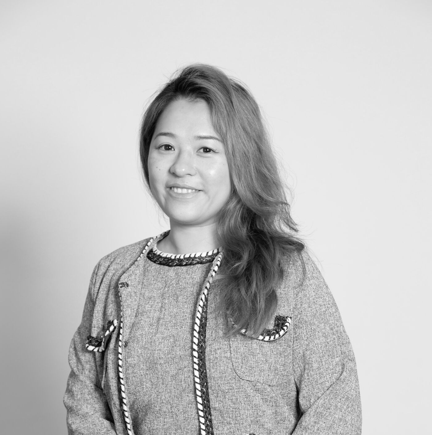 A monochrome, eye-level, medium shot depicts a woman with shoulder-length hair, wearing a tweed suit, seated with her hands clasped in her lap against a light-colored background.