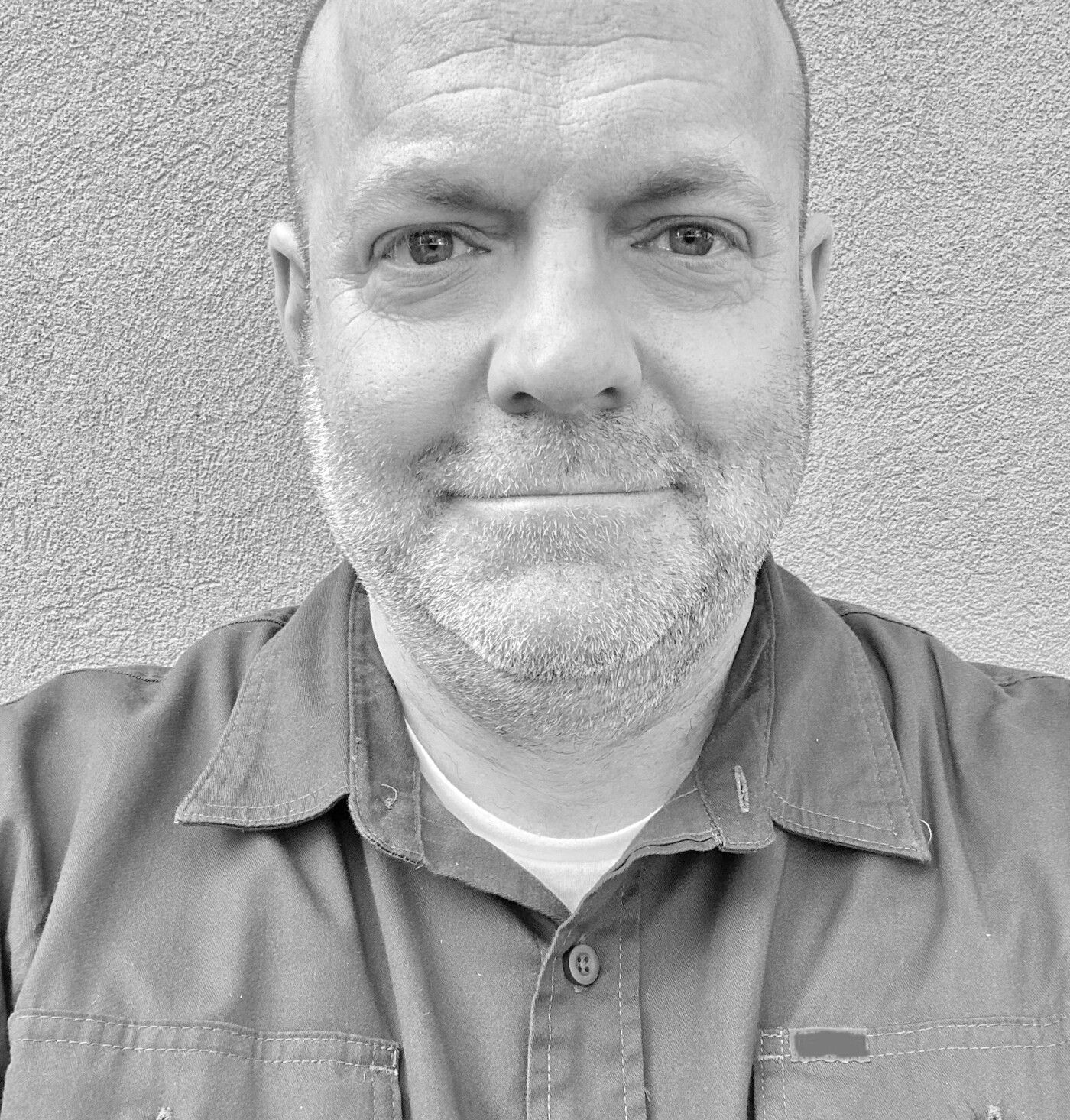 Close-up black and white shot of a man with short hair and a beard wearing a collared button-up shirt.