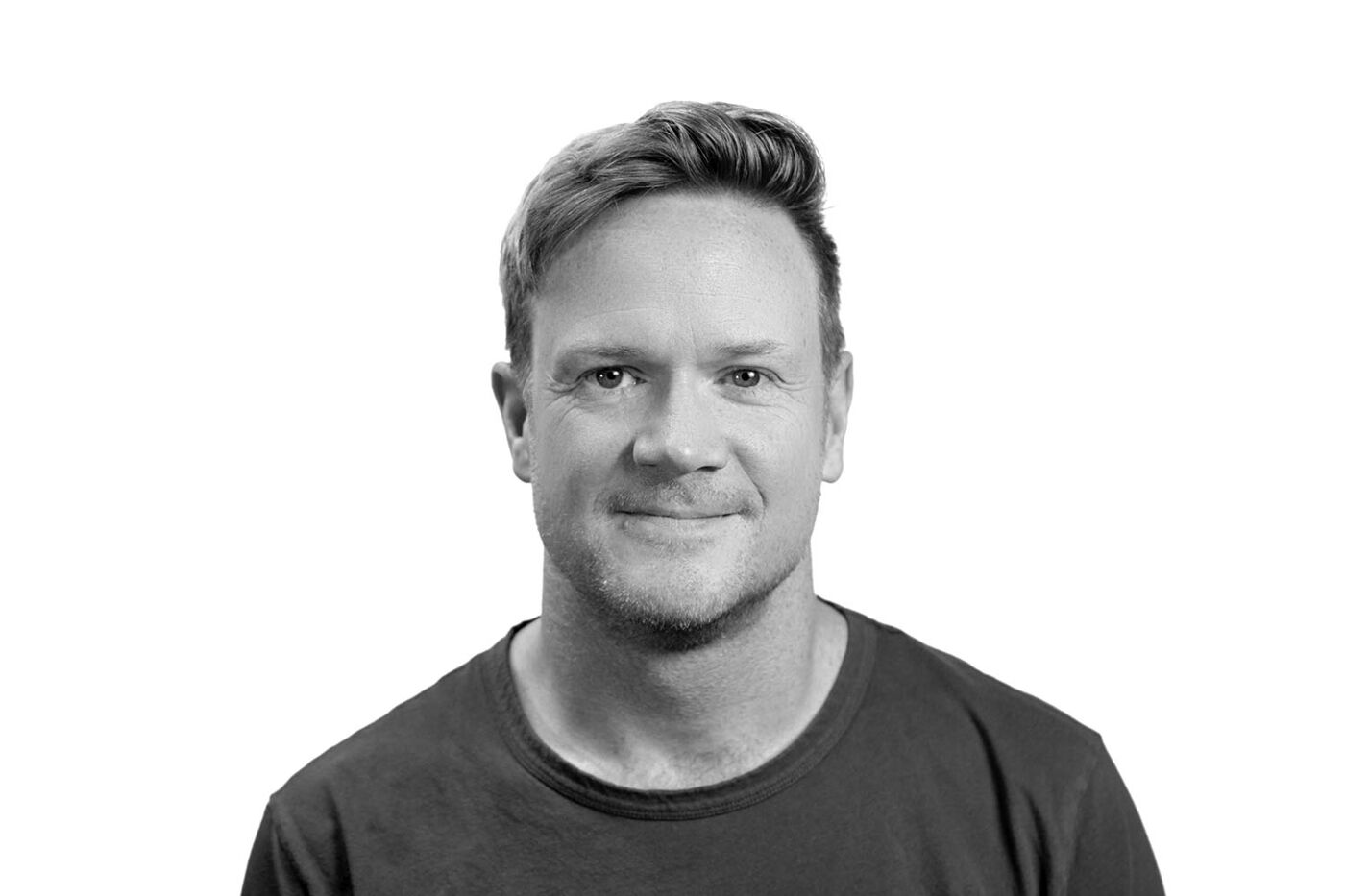 Close-up black and white shot of a man with short, slightly tousled hair, wearing a dark-colored crew-neck shirt, looking directly at the camera with a neutral expression.