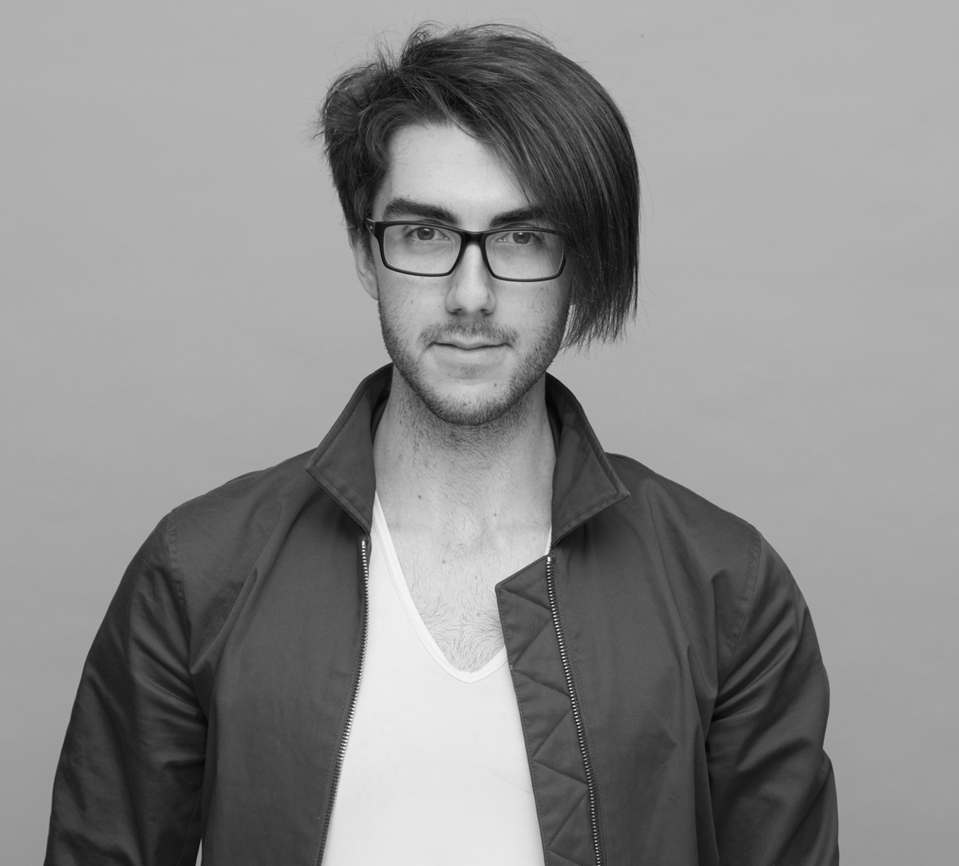 Close-up black and white shot of a young man with long hair, wearing glasses and a jacket, against a light gray background.
