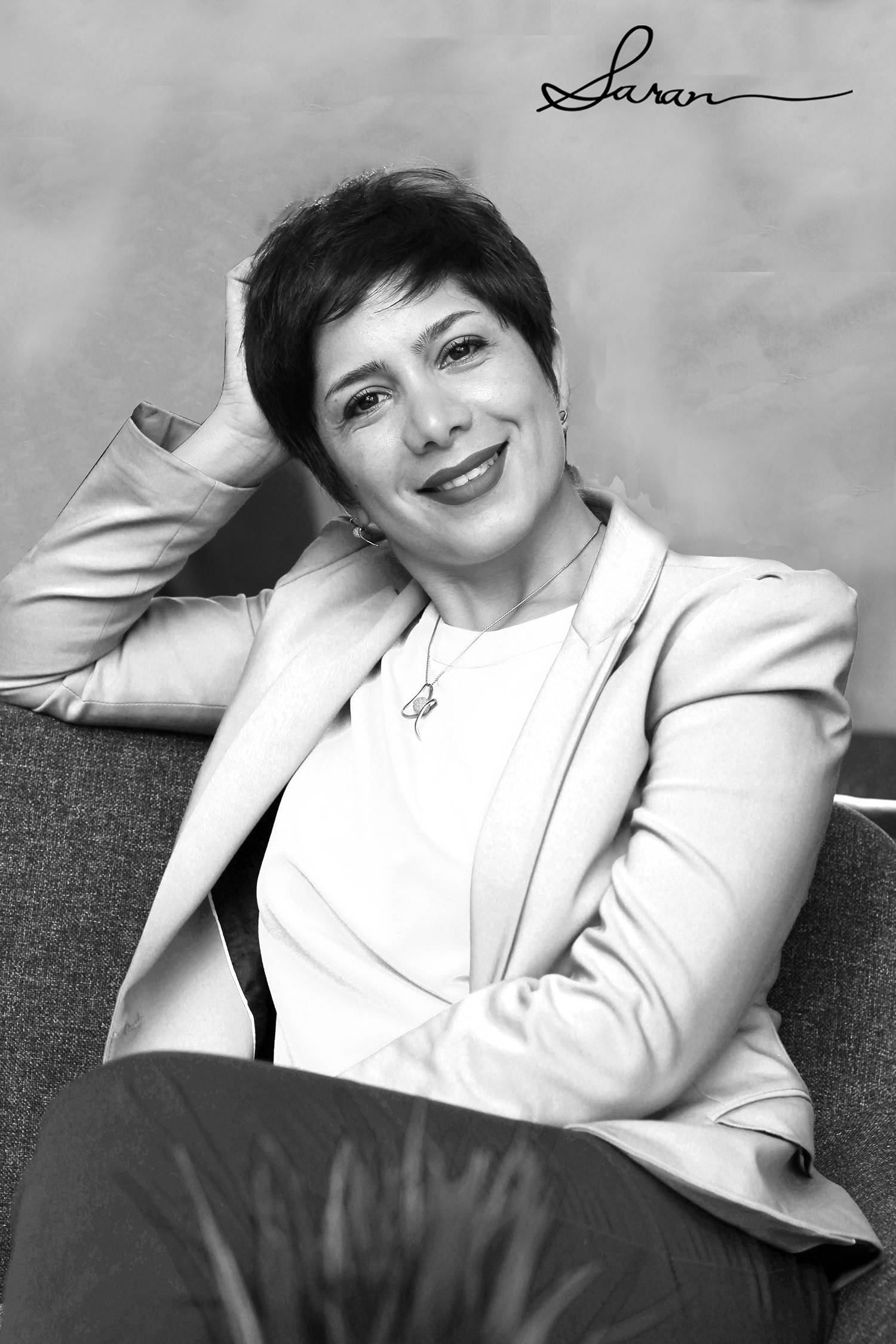 A black and white, medium shot of a woman named Saran, with short hair, wearing a light-colored blazer and a white shirt, sitting on a chair with a relaxed posture, her hand gently touching her hair.