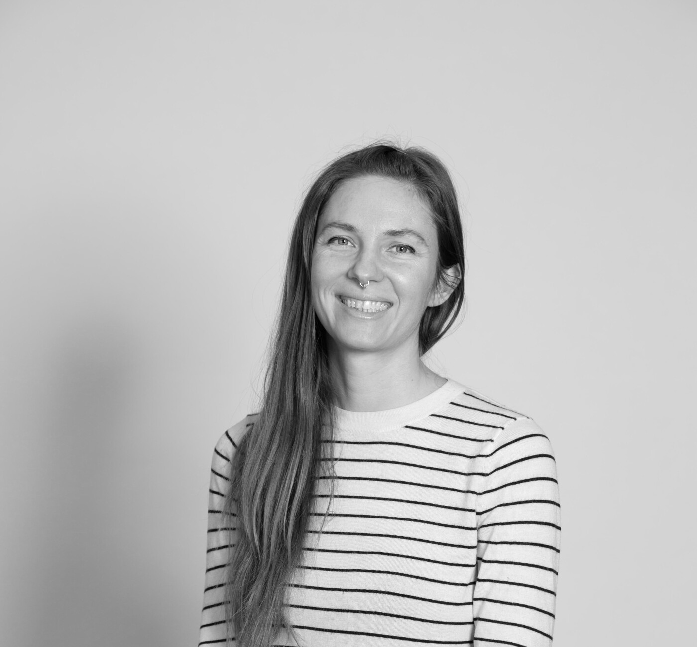 A black and white, eye-level, medium shot of a woman with long brown hair smiling while sitting on a stool, wearing a horizontally striped long-sleeve shirt and dark pants.