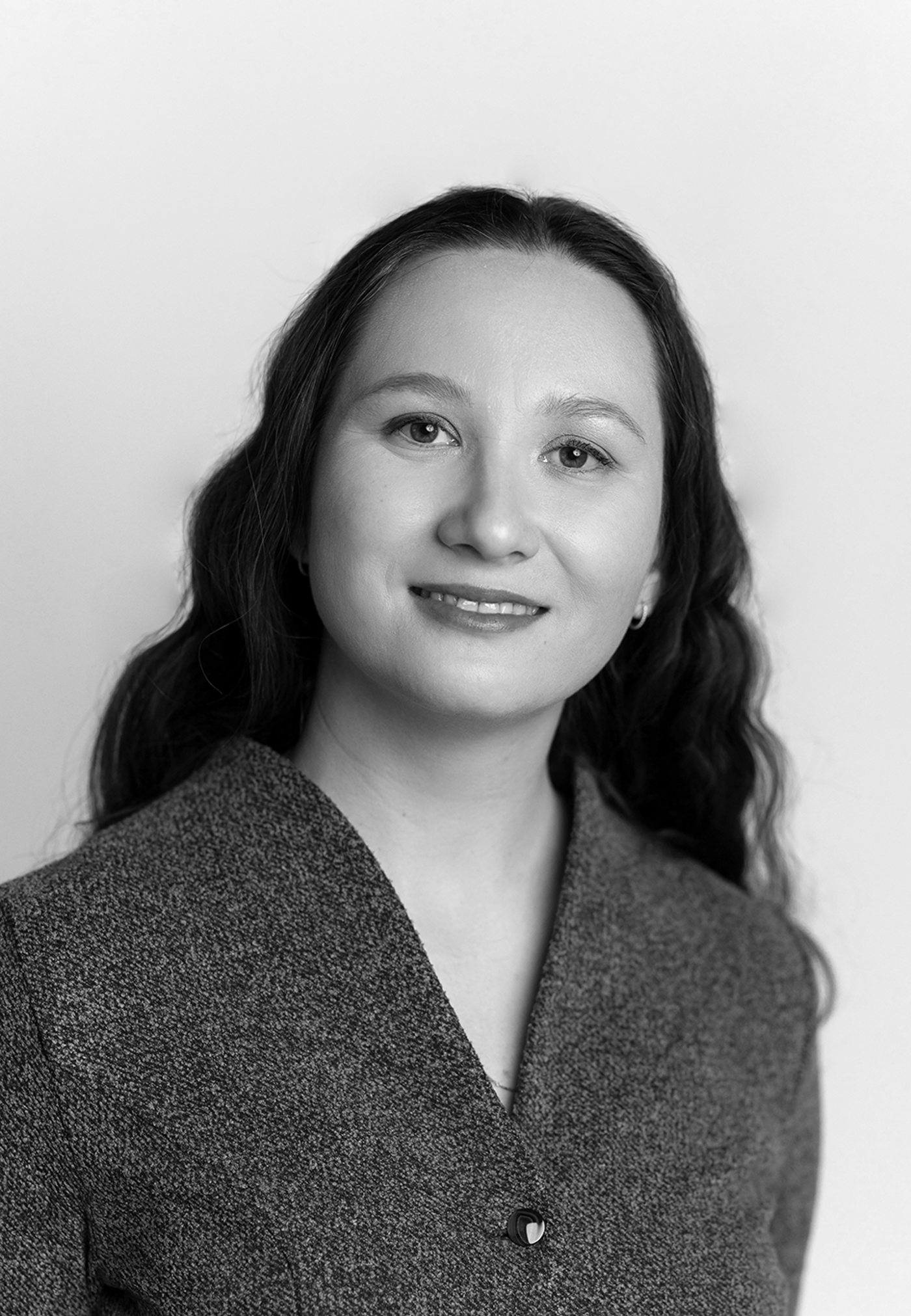 Close-up black and white portrait of a woman with long, wavy hair, wearing a textured v-neck jacket, smiling gently at the camera.