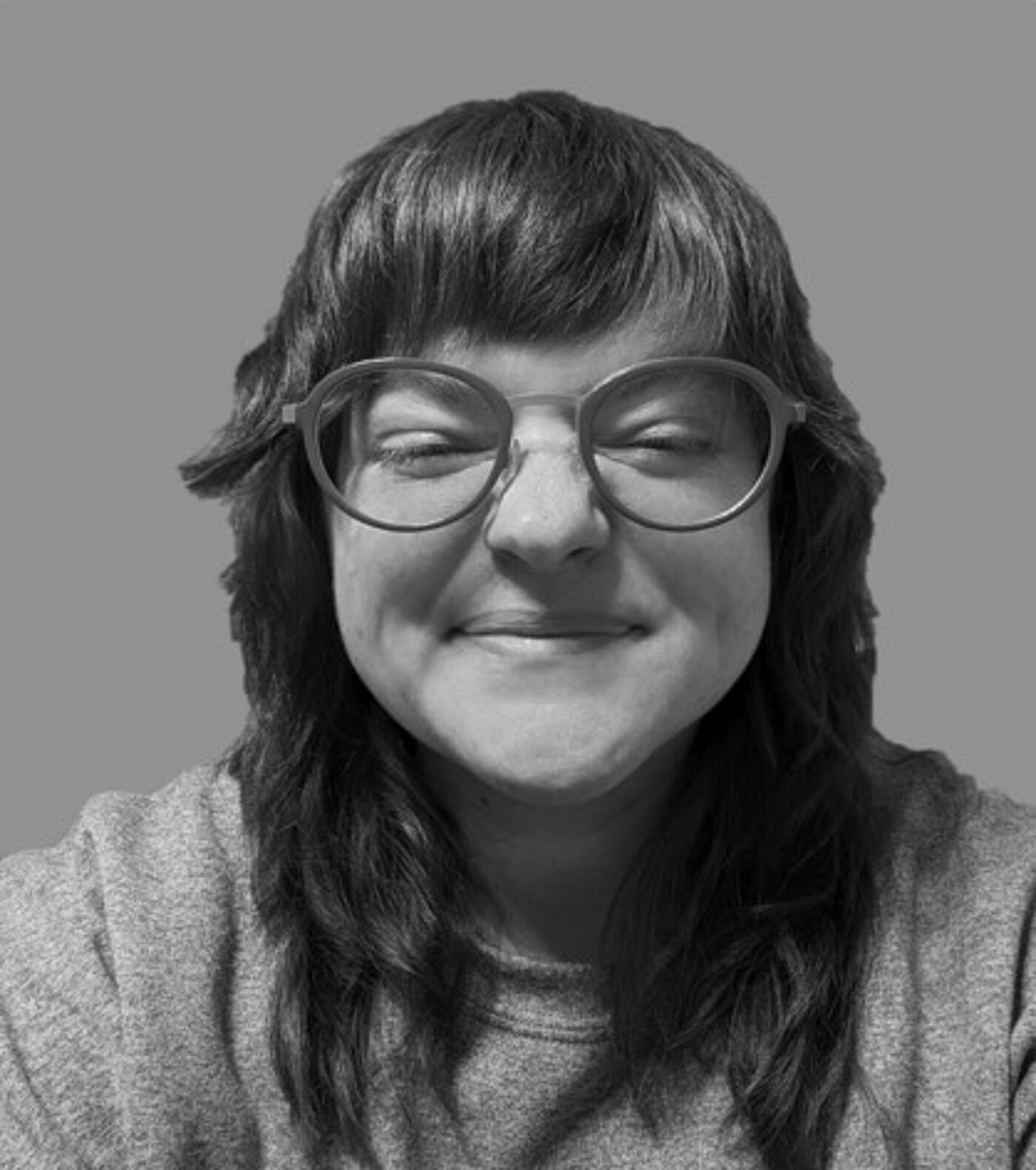 Close-up grayscale shot of a woman with long, dark hair and glasses, wearing a gray sweater, and smiling slightly.